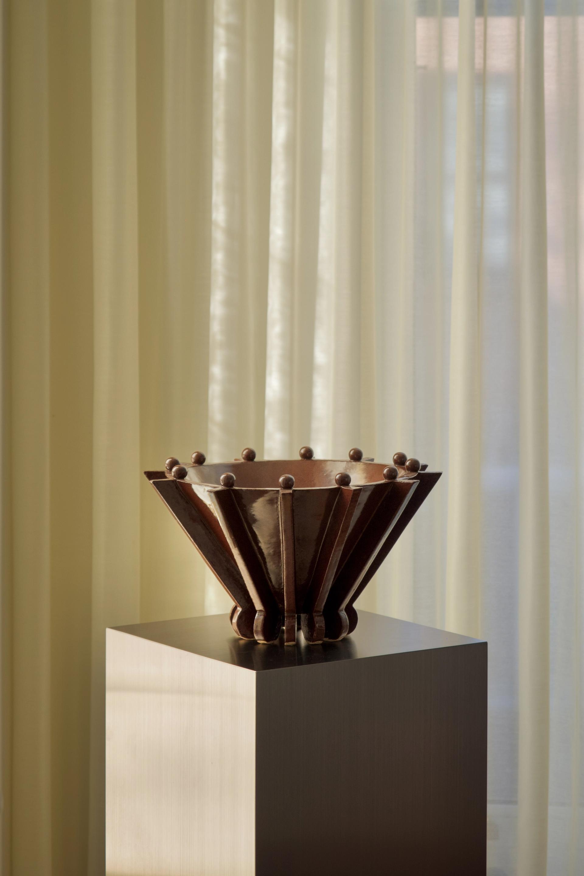 Brown ceramic vessel on a silver plinth with a backdrop of sheer curtains.