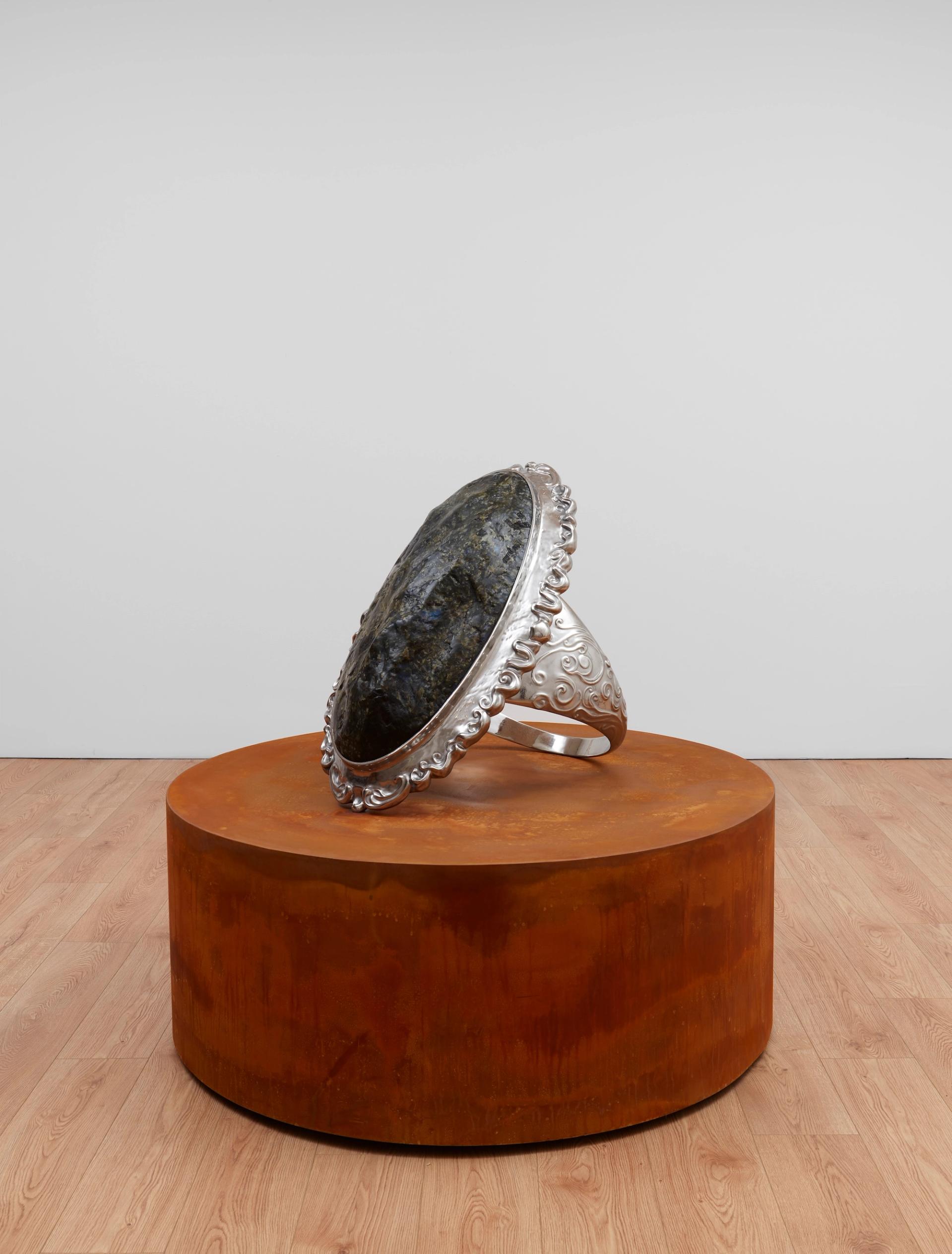 An oversized sculpted ring with an ornate silver setting and a dark stone, displayed upright on a rust-colored cylindrical plinth