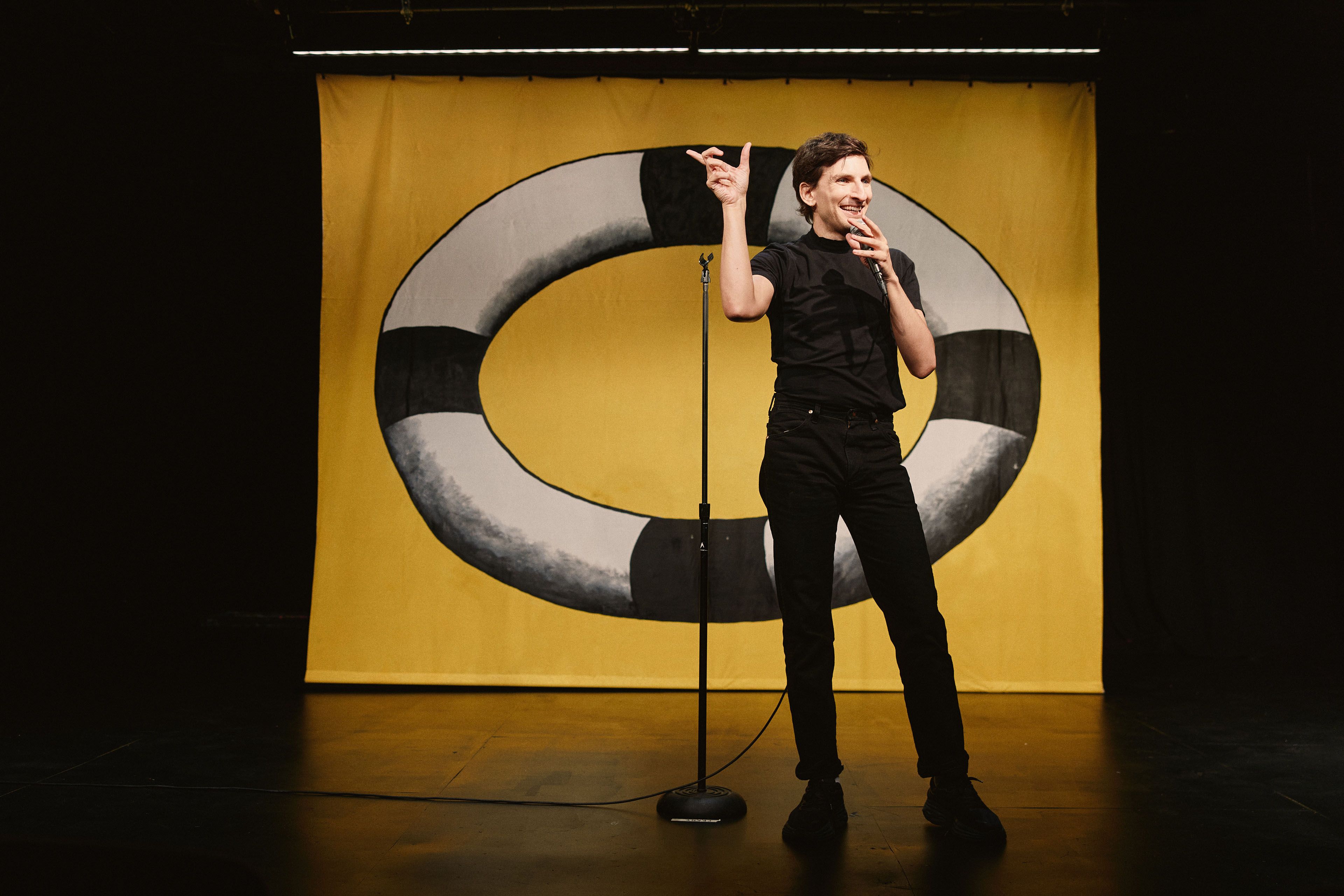 A performer in all black holds a microphone on a darkened stage, gesturing expressively in front of a yellow backdrop painted with a large black and white ring.