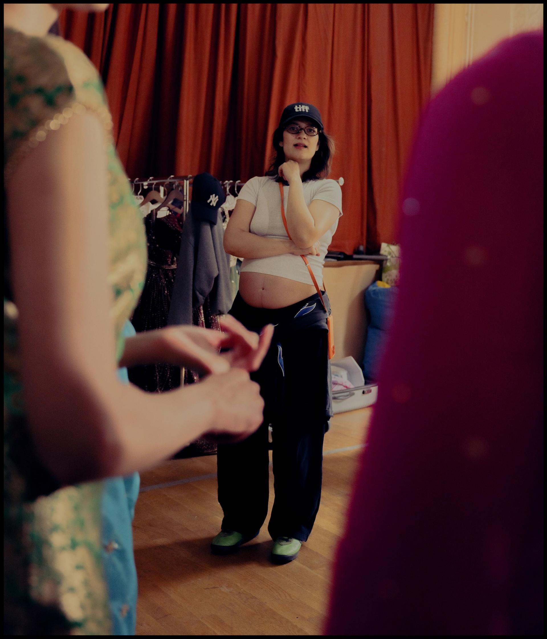 A woman in a cap and casual clothing observes performers in South Asian costume in a room with a clothing rack visible behind her.