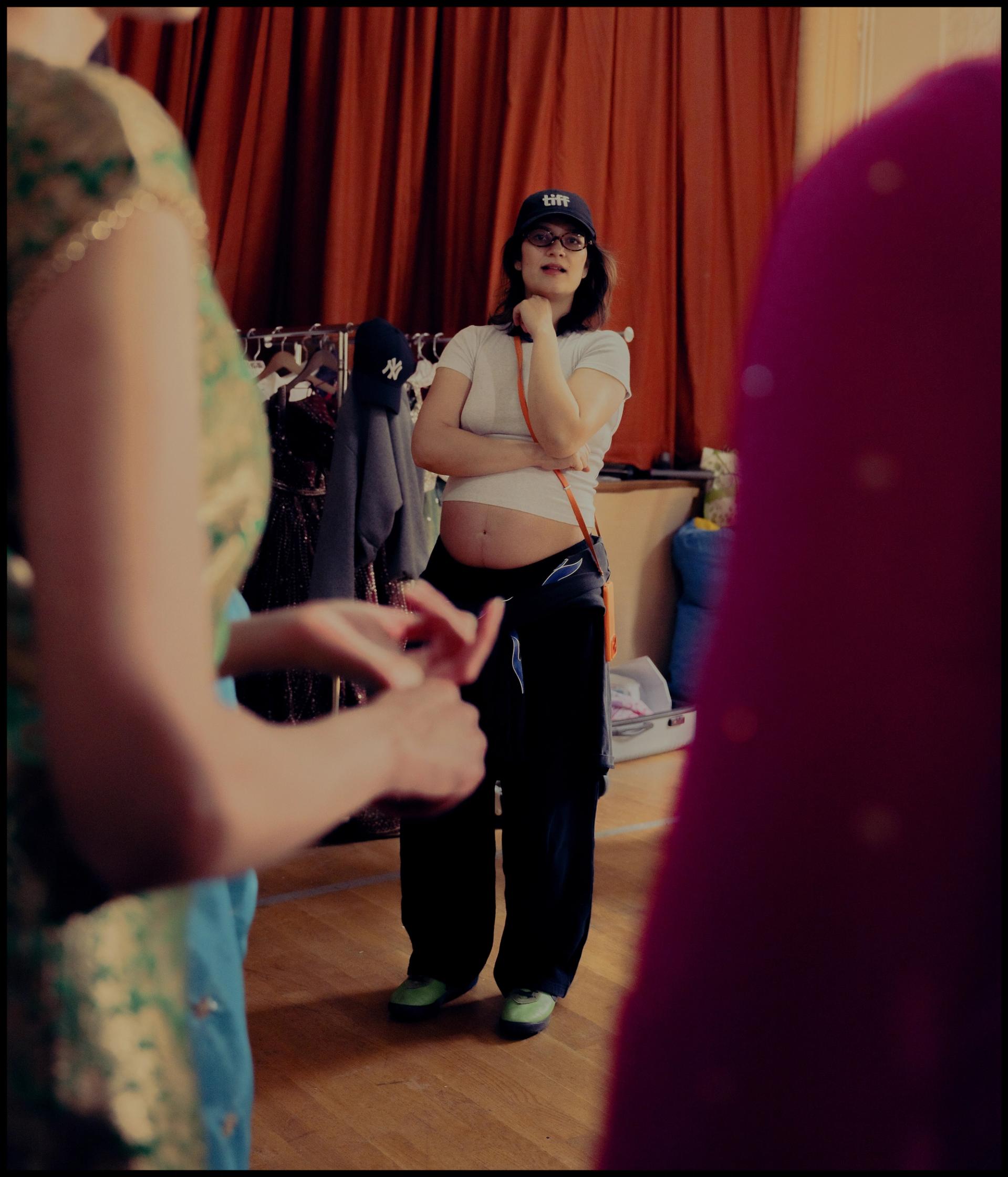 A woman in a cap and casual clothing observes performers in South Asian costume in a room with a clothing rack visible behind her.