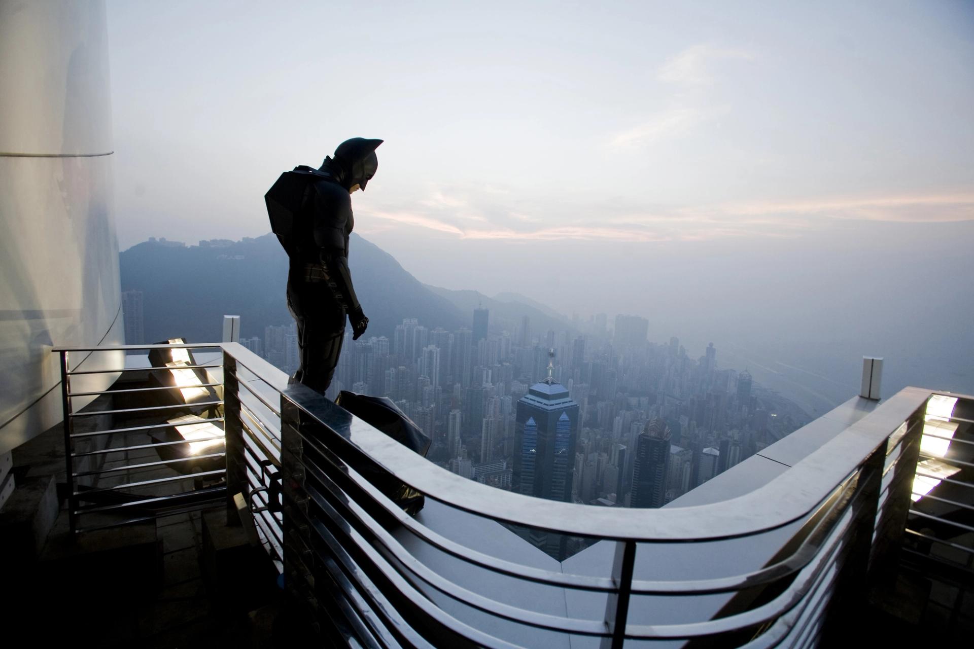 A figure in a black superhero costume stands on a high-rise ledge overlooking a dense city skyline shrouded in haze