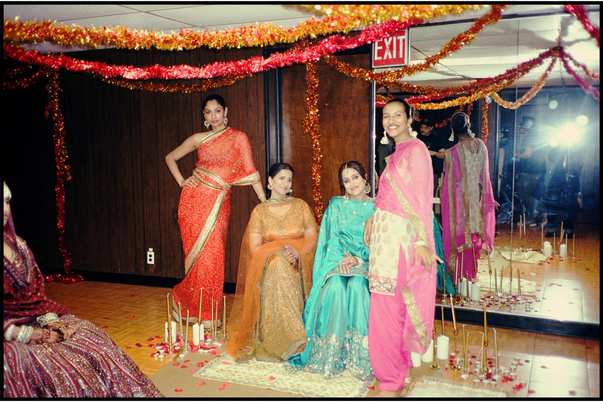 Four performers in South Asian festive dress pose on a decorated film set strung with gold and red tinsel garlands, with candles and rose petals on the floor and a mirrored wall reflecting the crew behind them