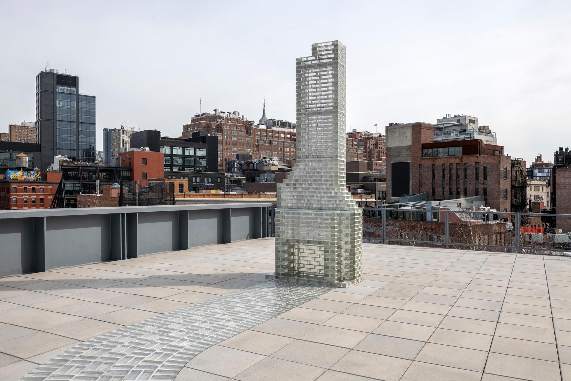 A tall translucent glass brick chimney sculpture stands on a rooftop terrace, set against a city skyline under an overcast sky