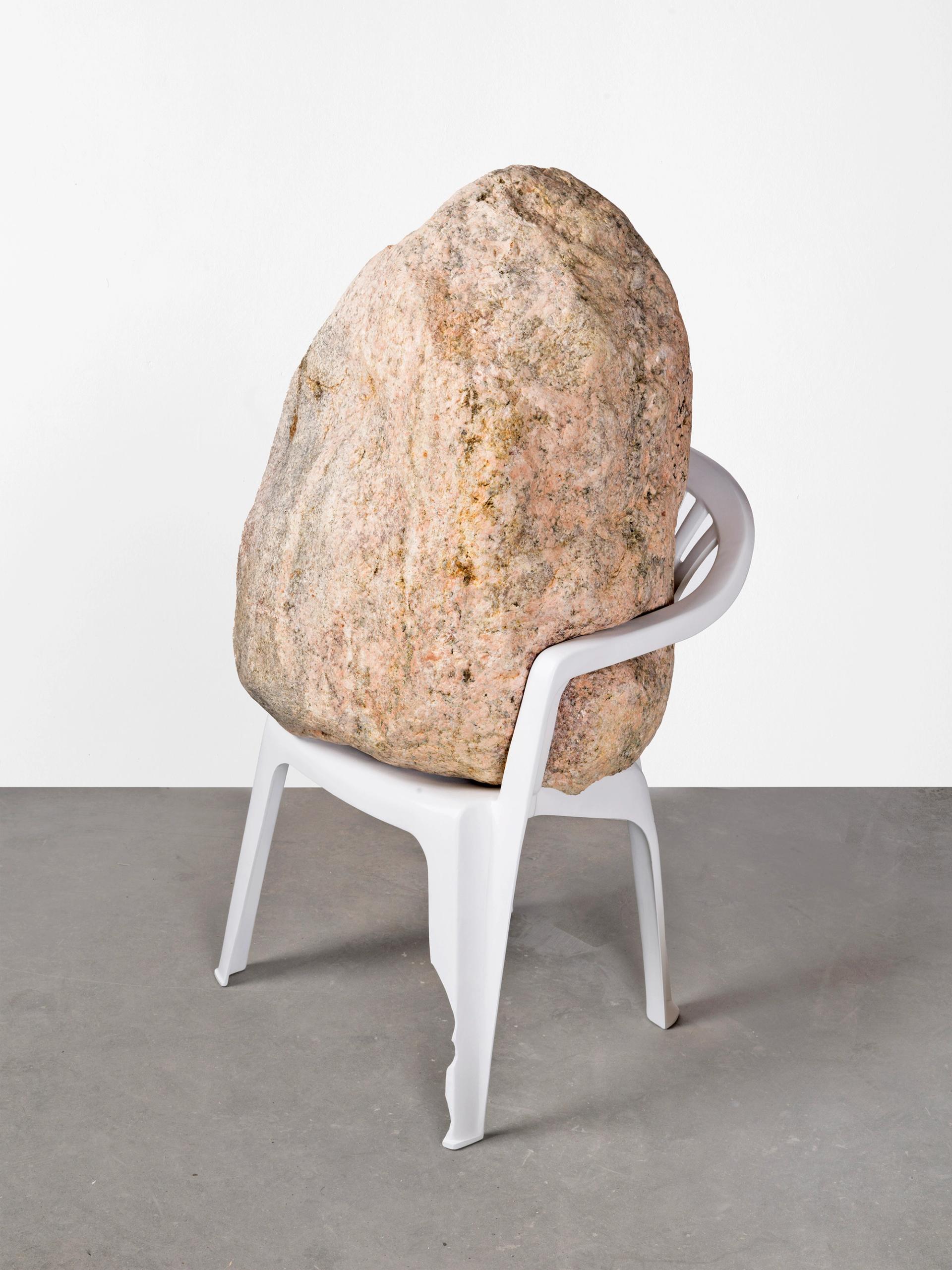 Large stone balanced inside white chair as sculptural installation against gallery wall.