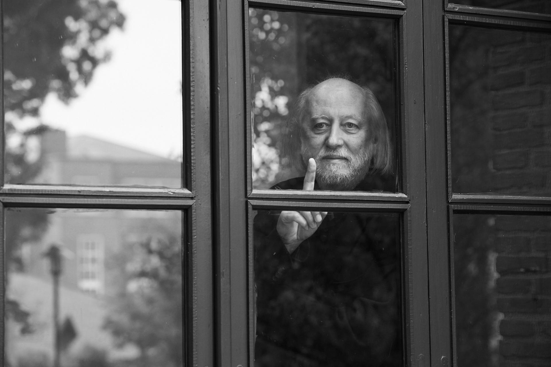 Black-and-white photograph of a man standing behind a window frame and holding up his right index finger.
