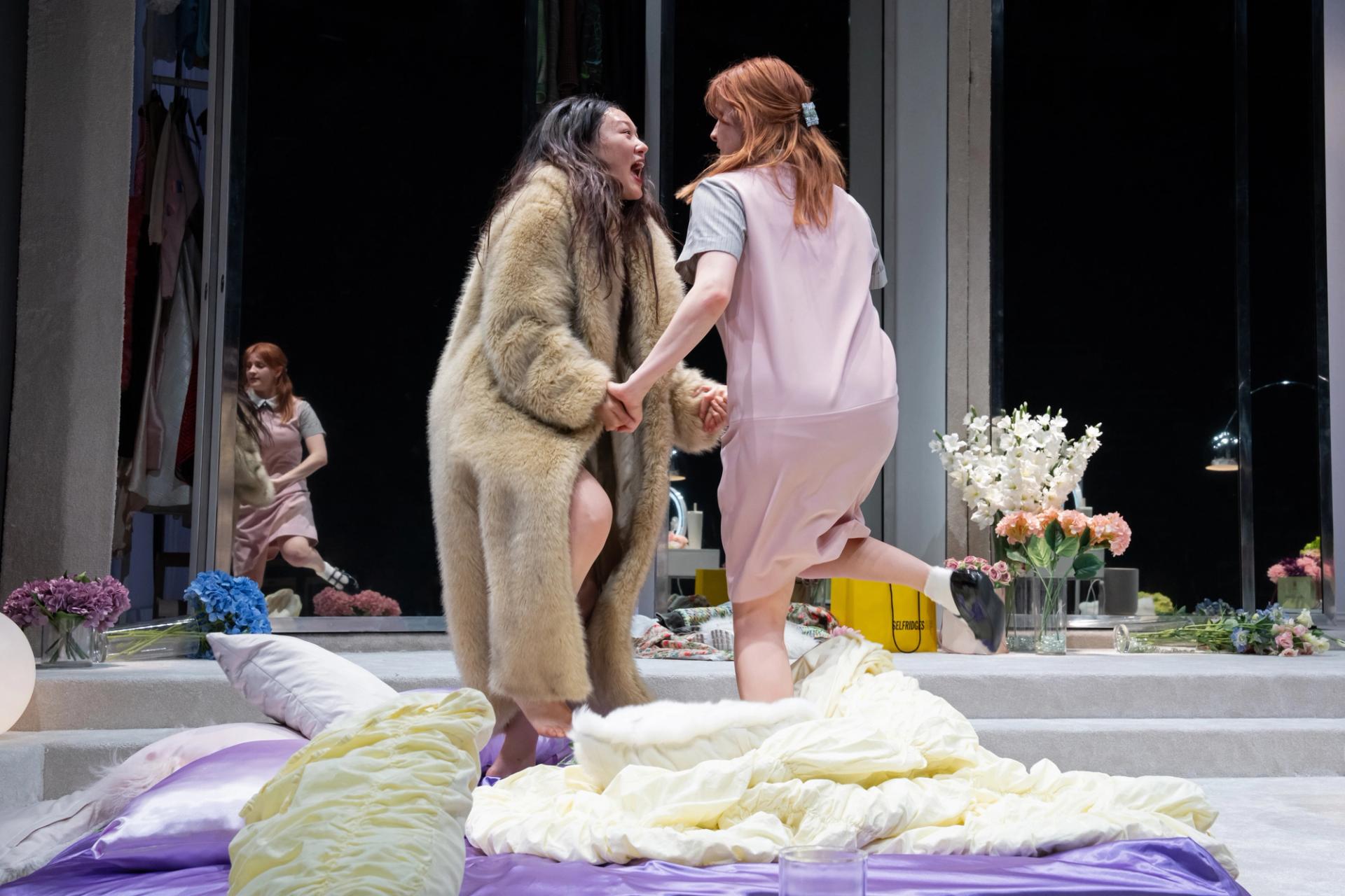 Two performers clasp hands amid rumpled bedding and flowers on a stage set.