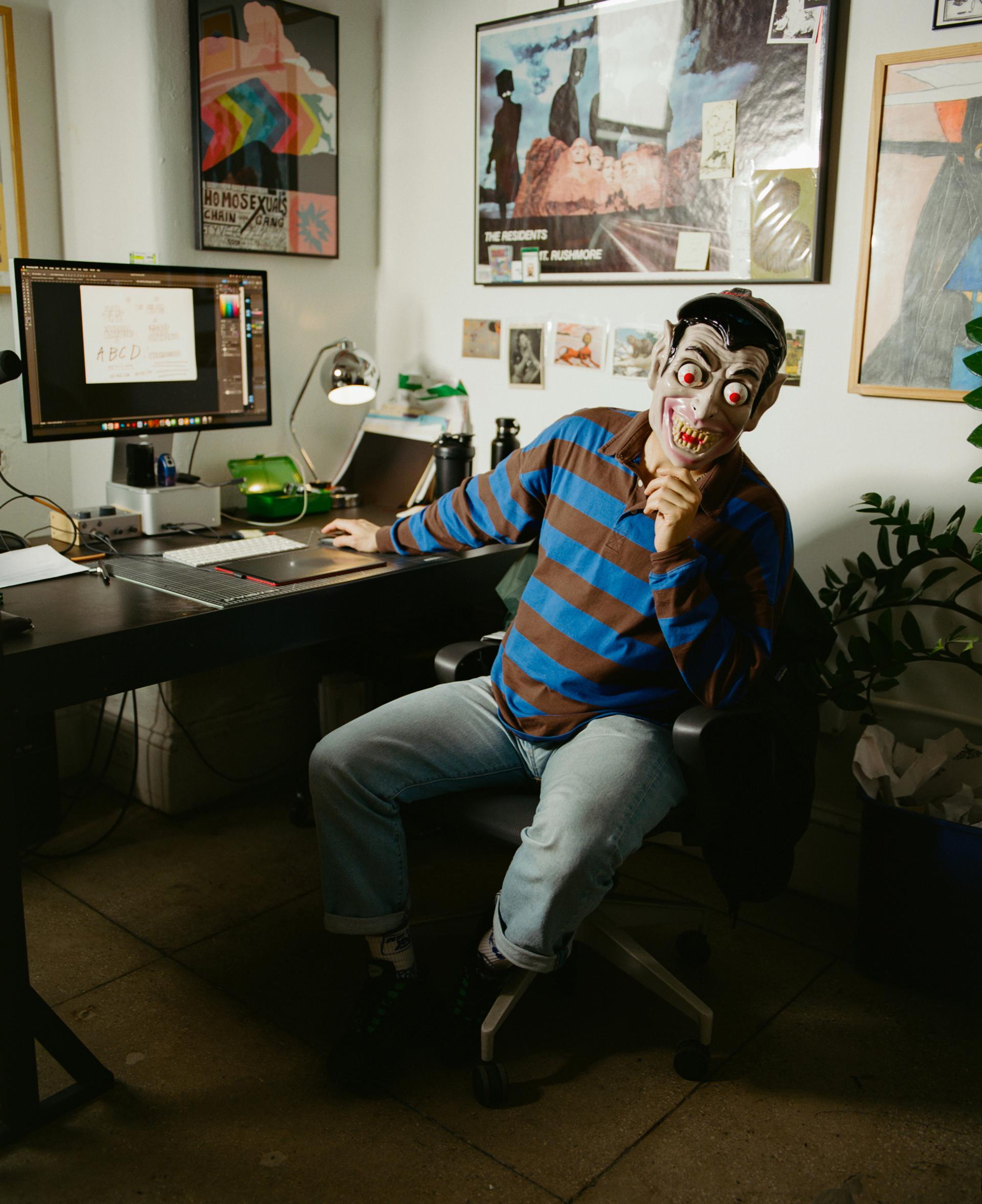 A man in a brown and blue striped shirt sits in a desk chair holding a mask with bulging eyes and jagged teeth in front of his face.