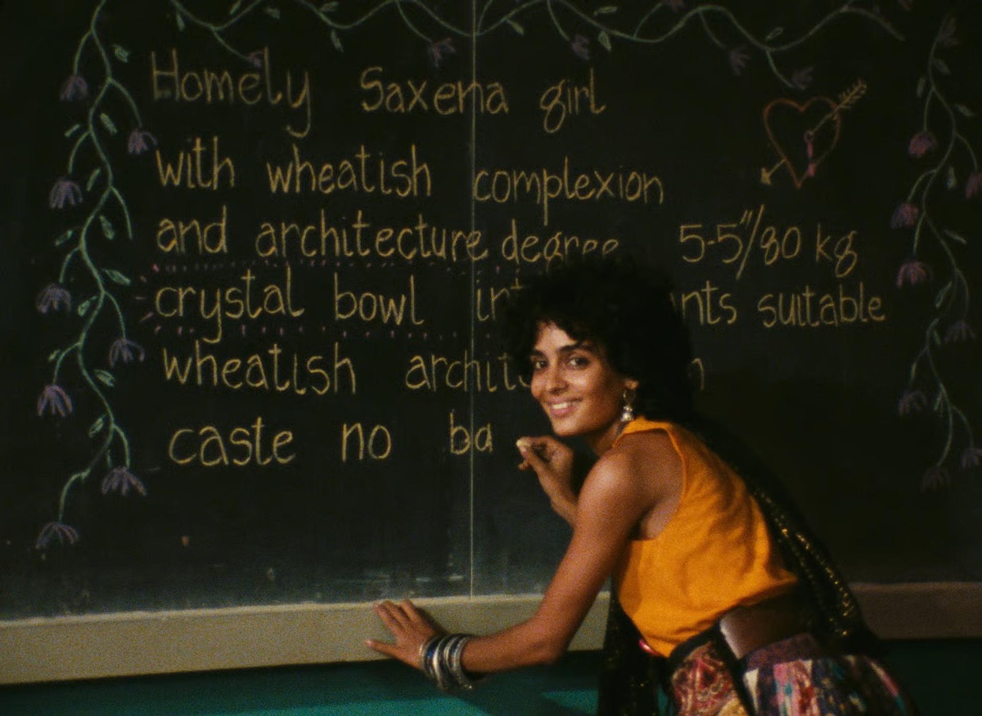 A smiling woman in an orange top crouched beside a chalkboard, partially covering handwritten text