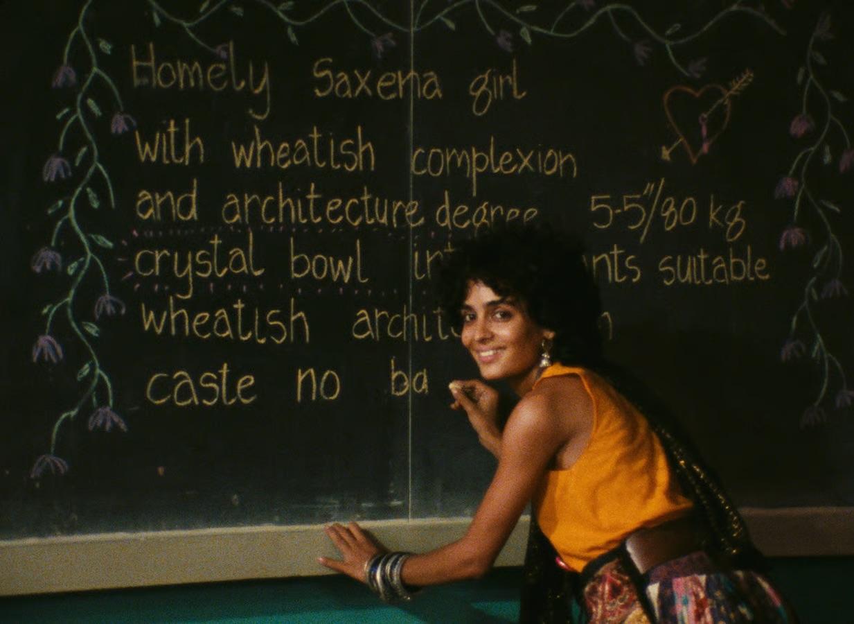 A smiling woman in an orange top crouched beside a chalkboard, partially covering handwritten text