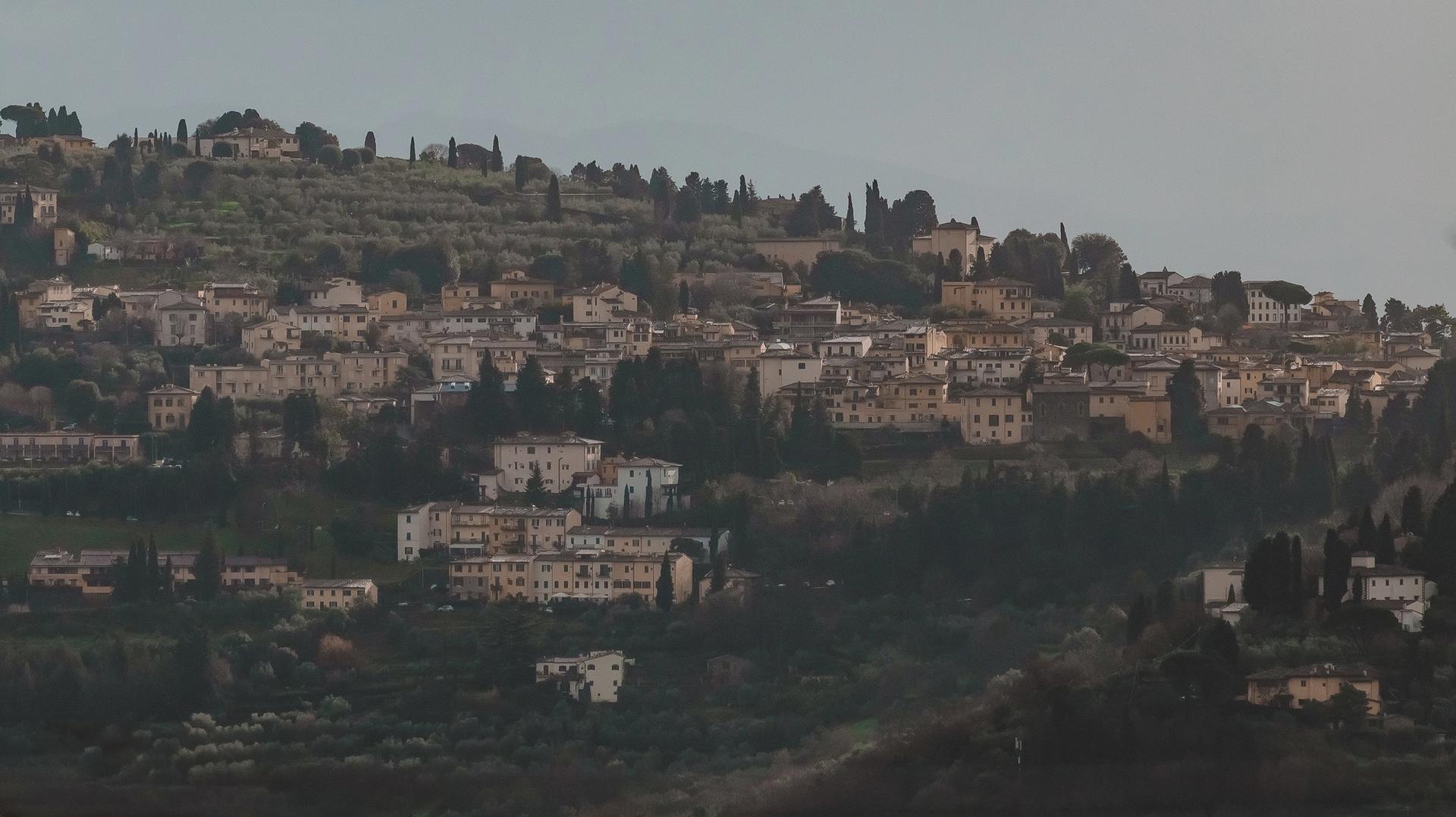 A hillside town with stone buildings and cypress trees under an overcast sky, viewed from a distance.