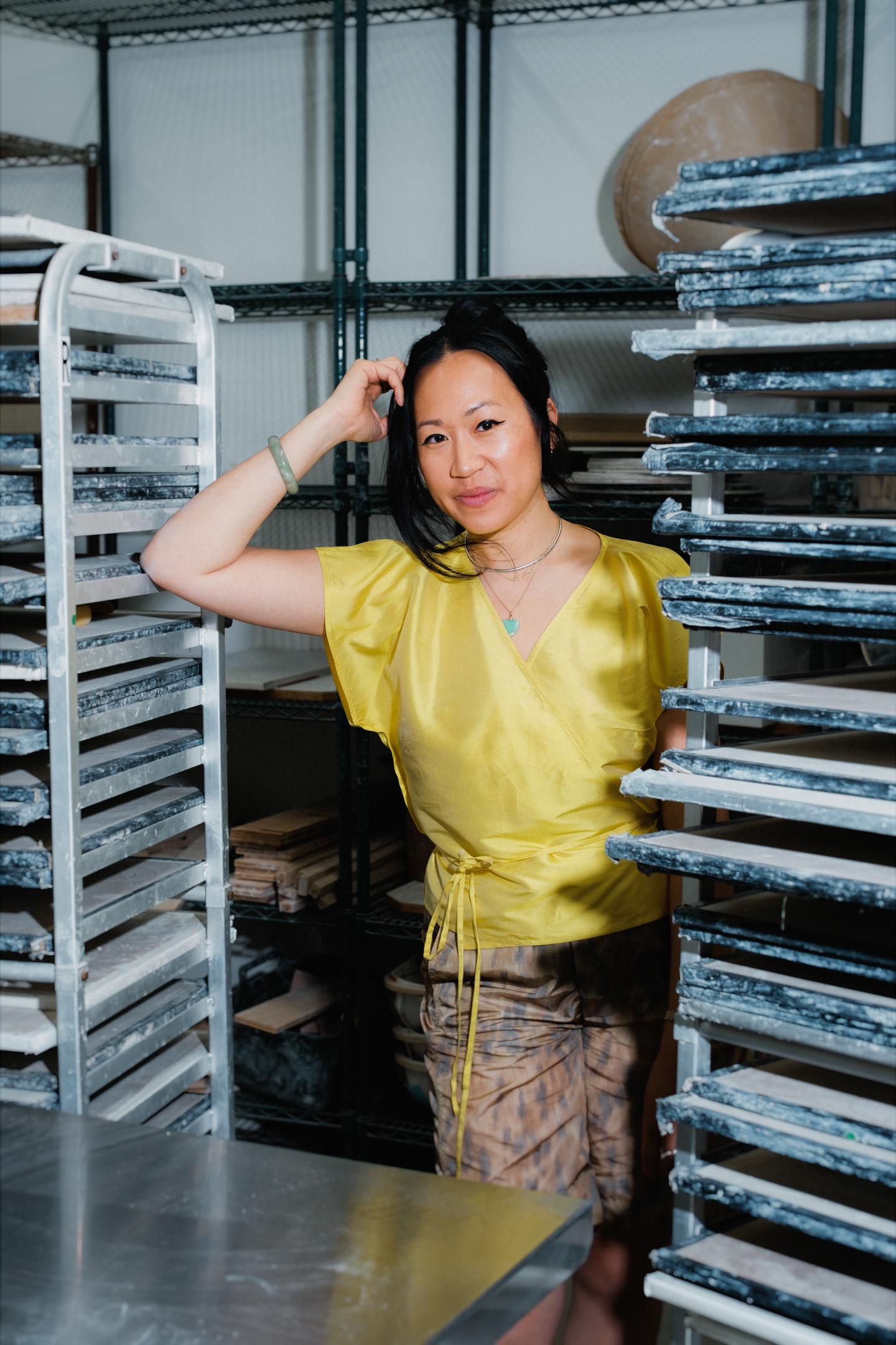 A woman in a yellow wrap top standing between tall metal racks loaded with drying ceramic slabs, one hand resting against her head