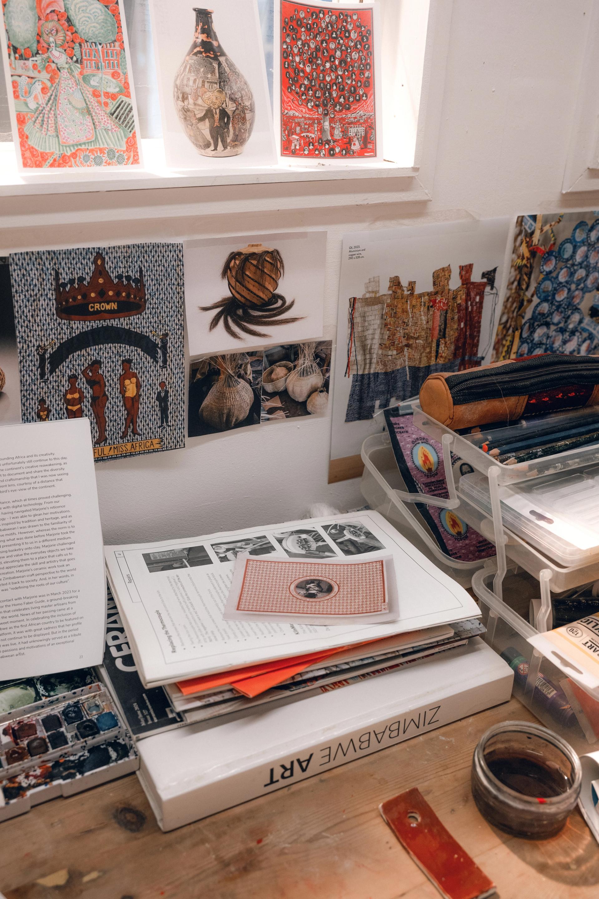 A cluttered desk with art books including one titled Zimbabwe Art, open magazines, reference images of woven baskets, and colorful prints displayed on a windowsill behind.