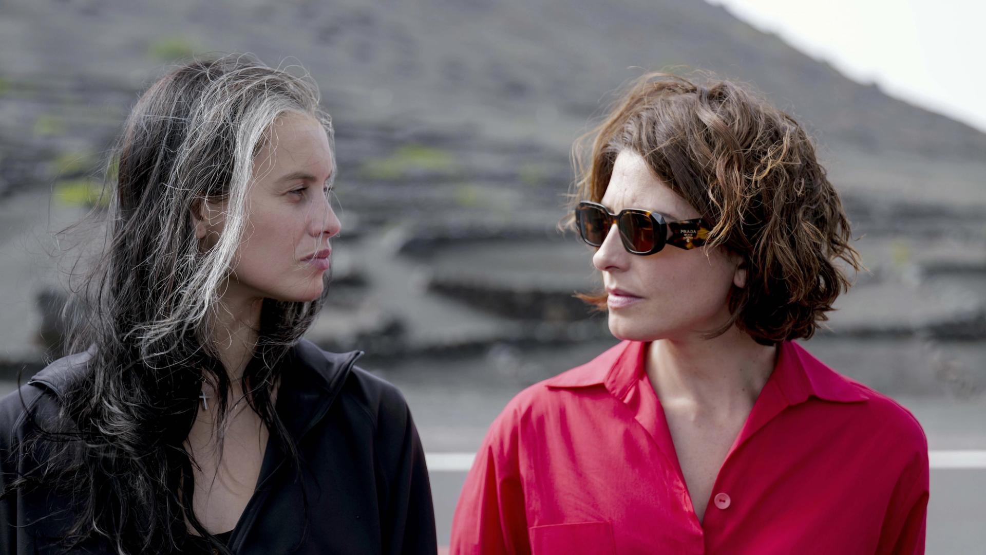 Two women standing outdoors in volcanic landscape, facing one another in black and bright red clothing.