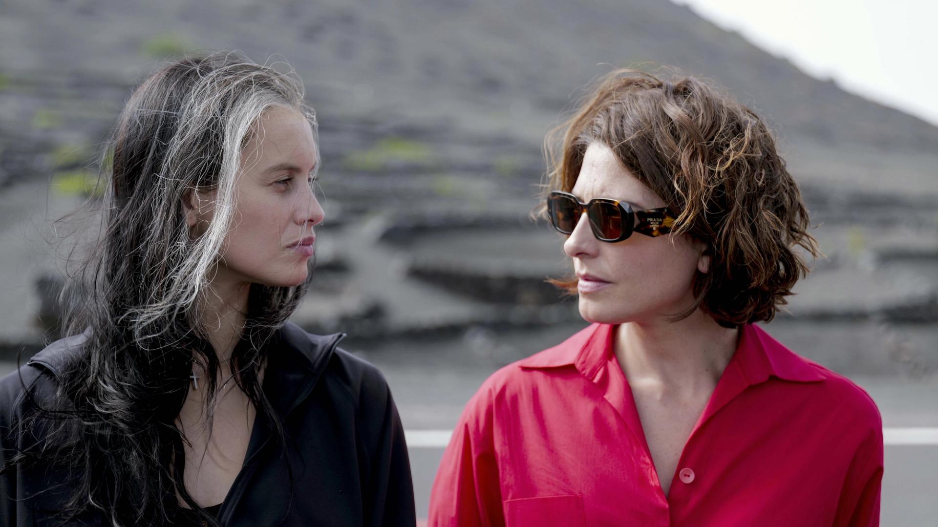 Two women standing outdoors in volcanic landscape, facing one another in black and bright red clothing.