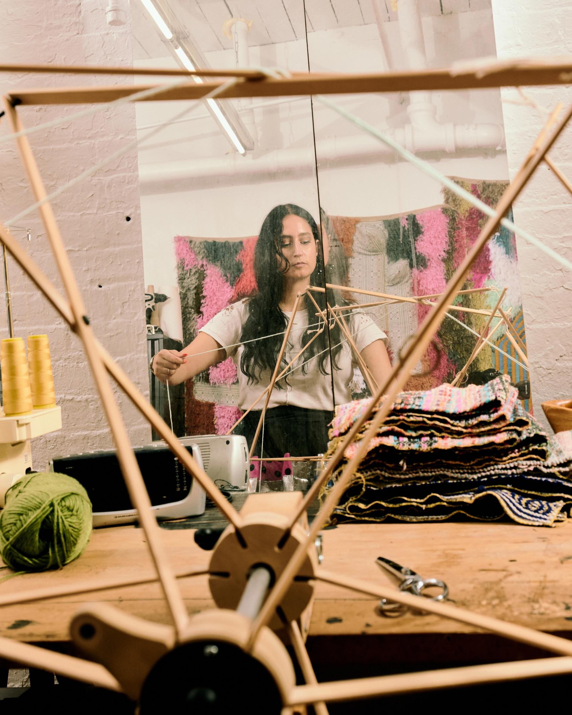 A woman winding thread on a wooden yarn swift, seen through the crossed wooden arms of the swift in the foreground, with colorful woven rugs, yarn spools, and fabric samples surrounding her workspace