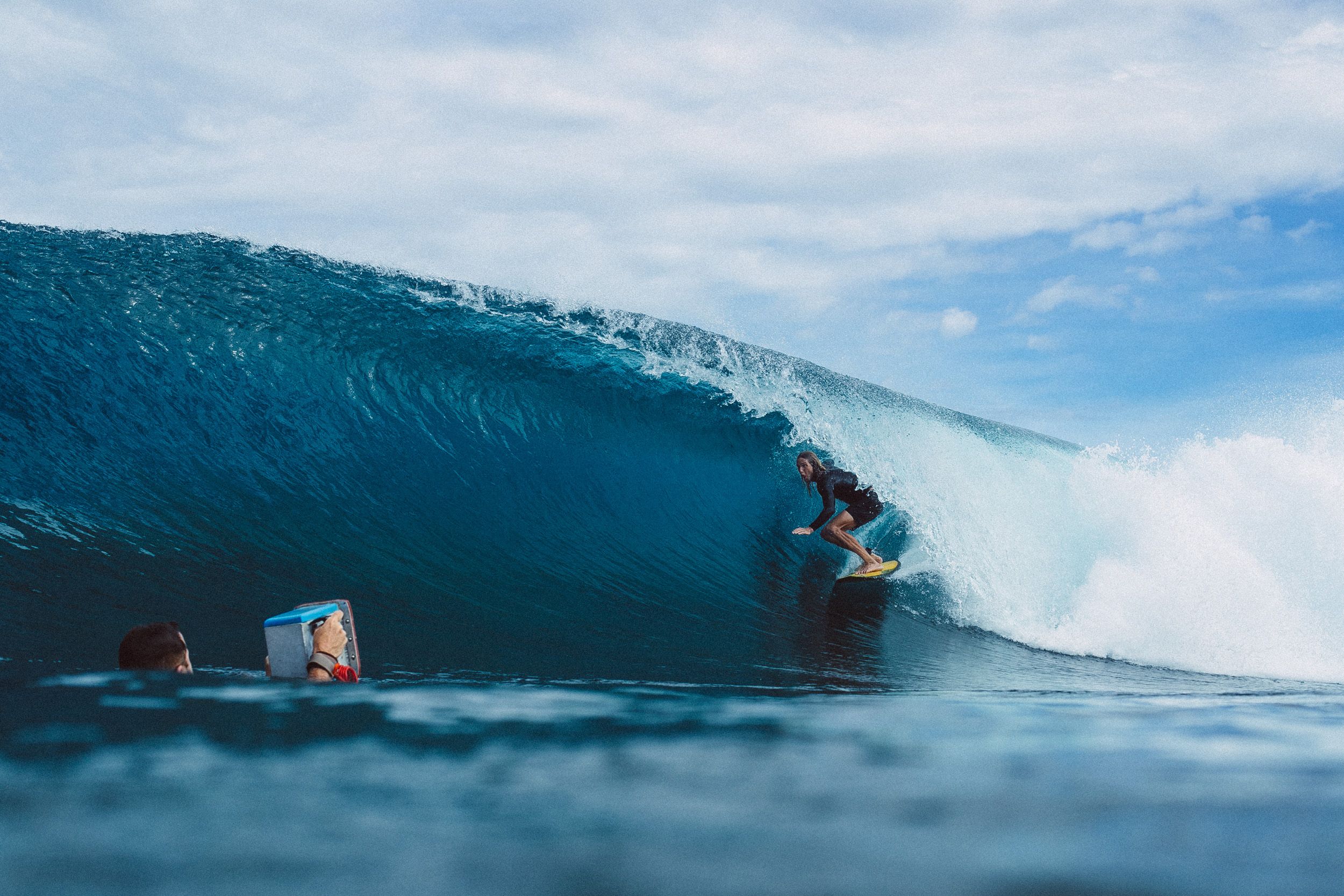 a surfer crouched inside a large barreling wave on a yellow surfboard, with a photographer floating in the foreground holding an underwater camera housing