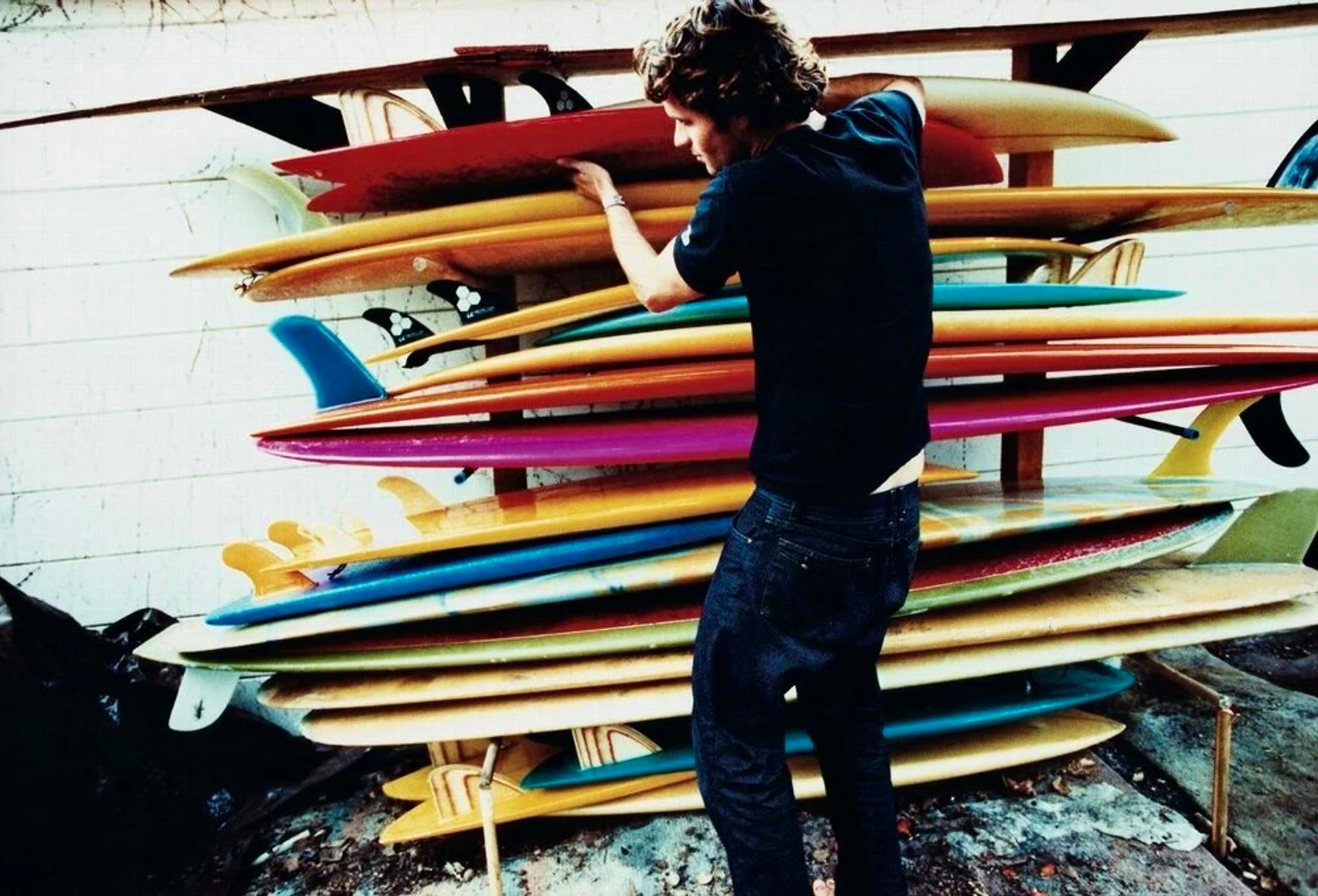A man reaching up to a tall rack densely packed with multicolored surfboards in red, yellow, blue, green, and pink, stacked horizontally against a white wall.
