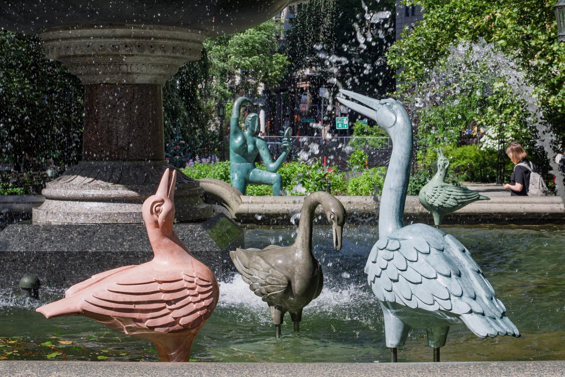 Several sculpted birds of various colors stand in the basin of a large public fountain, with a Cubist-inspired statue of a woman looking in a hand mirror visible in the background.