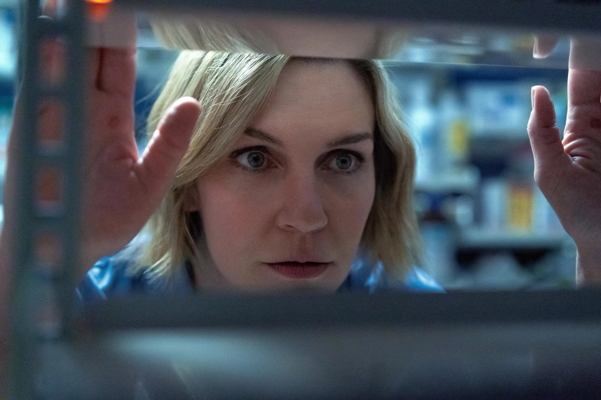A wide-eyed blond woman peering through a narrow gap between shelves, her face tightly framed with a hand raised near her hair, in a dim, blue-lit interior