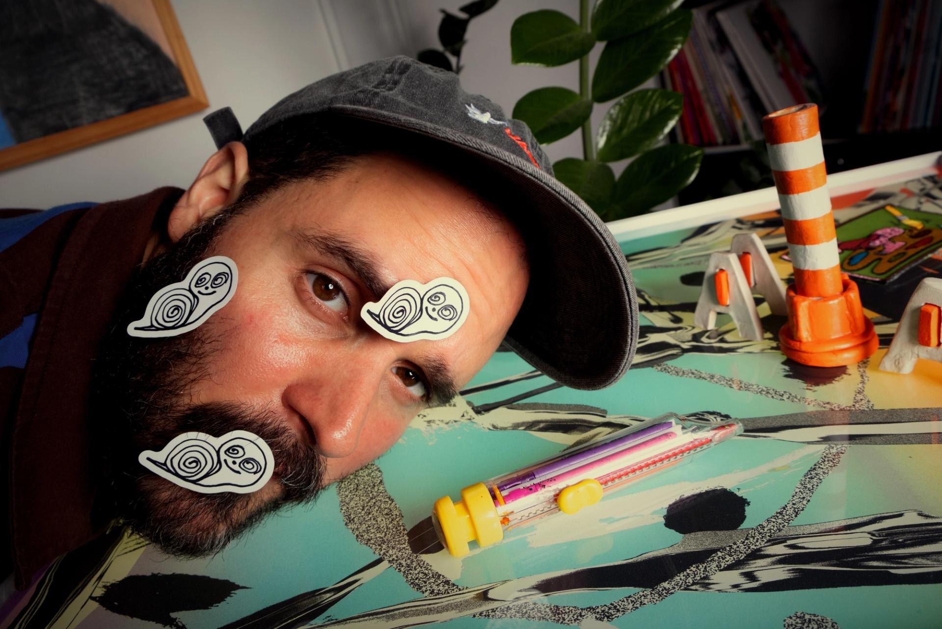 Man with a beard wearing a cap rests their head on a painted surface, with small cartoon snail stickers on their face. A box cutter and small striped sculpture sit nearby, with a plant and books in the background.