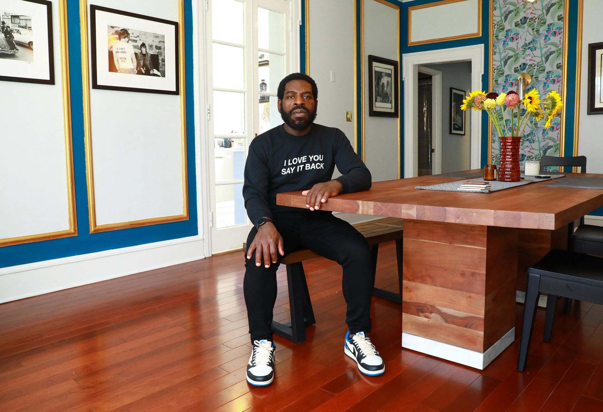 Color photo of a bearded man in a black sweatshirt reading "I love you, say it back" seated at a wooden table in room with framed black-and-white photos, patterned wallpaper, and a vase of sunflowers.