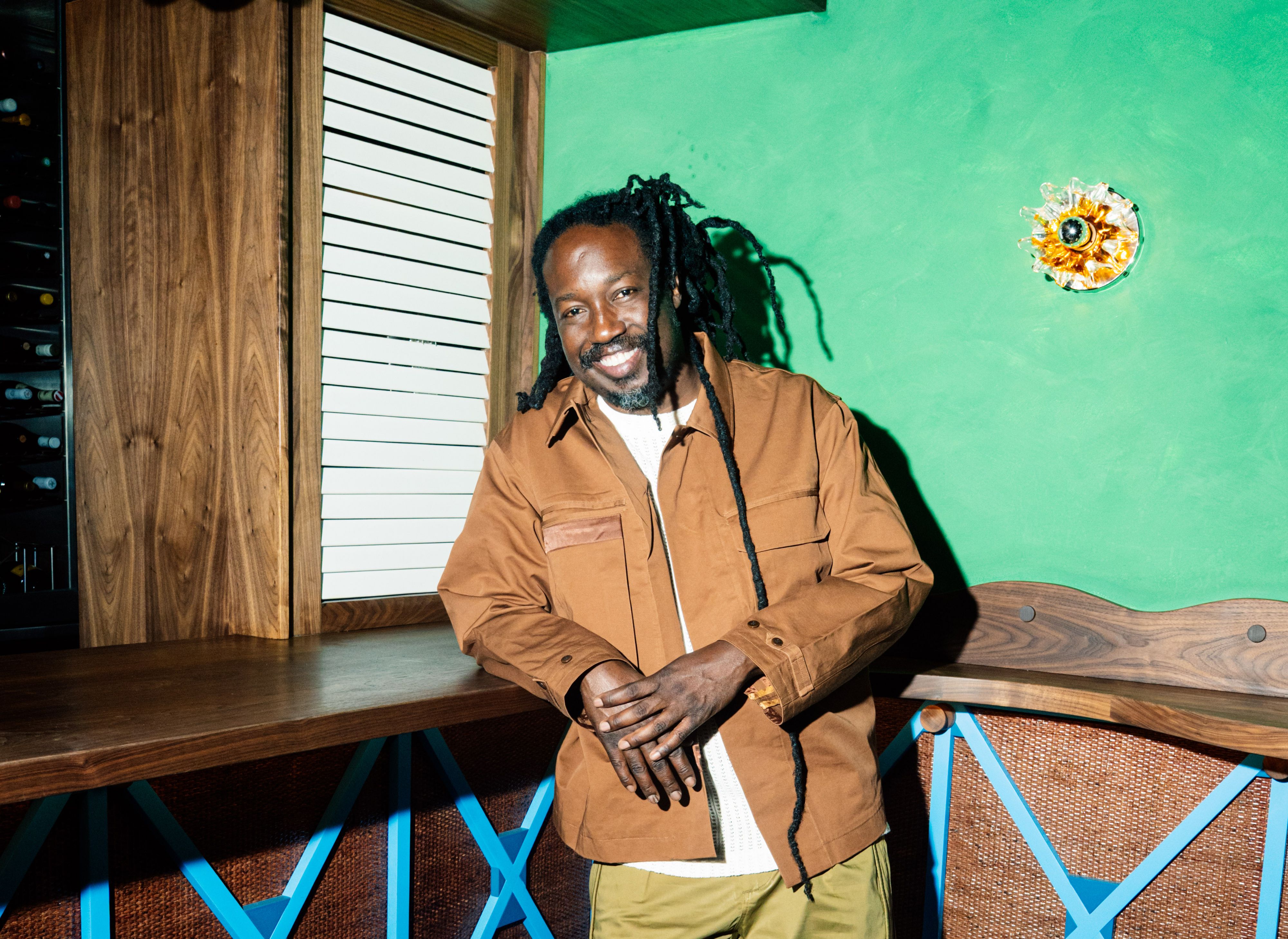 A man with dreadlocks wearing a tan jacket and white shirt leans against a wooden bar in front of a bright green wall.