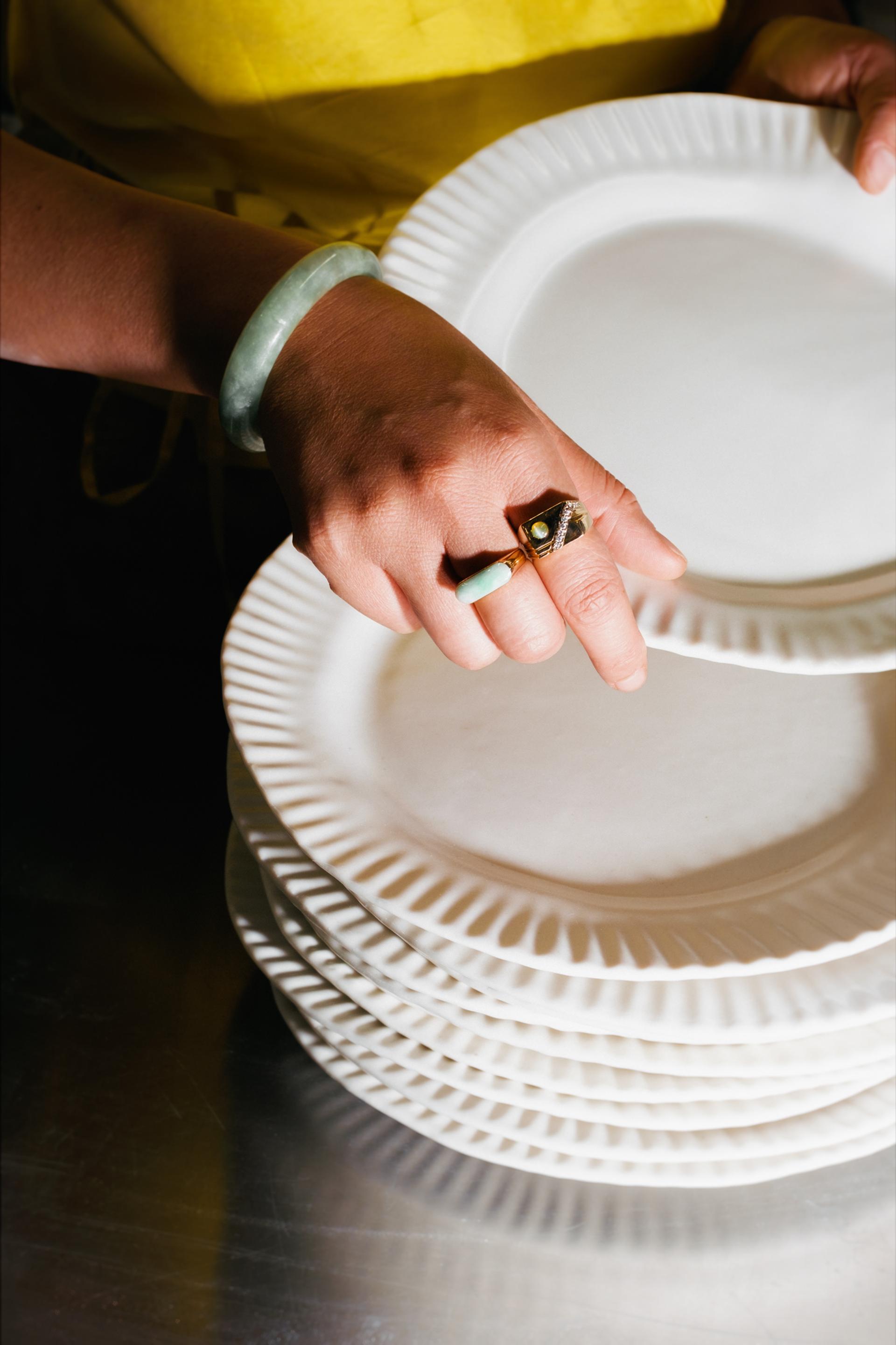 Close-up color photo of a hand wearing a jade bangle and gold ring lifting a fluted white plate from a stack