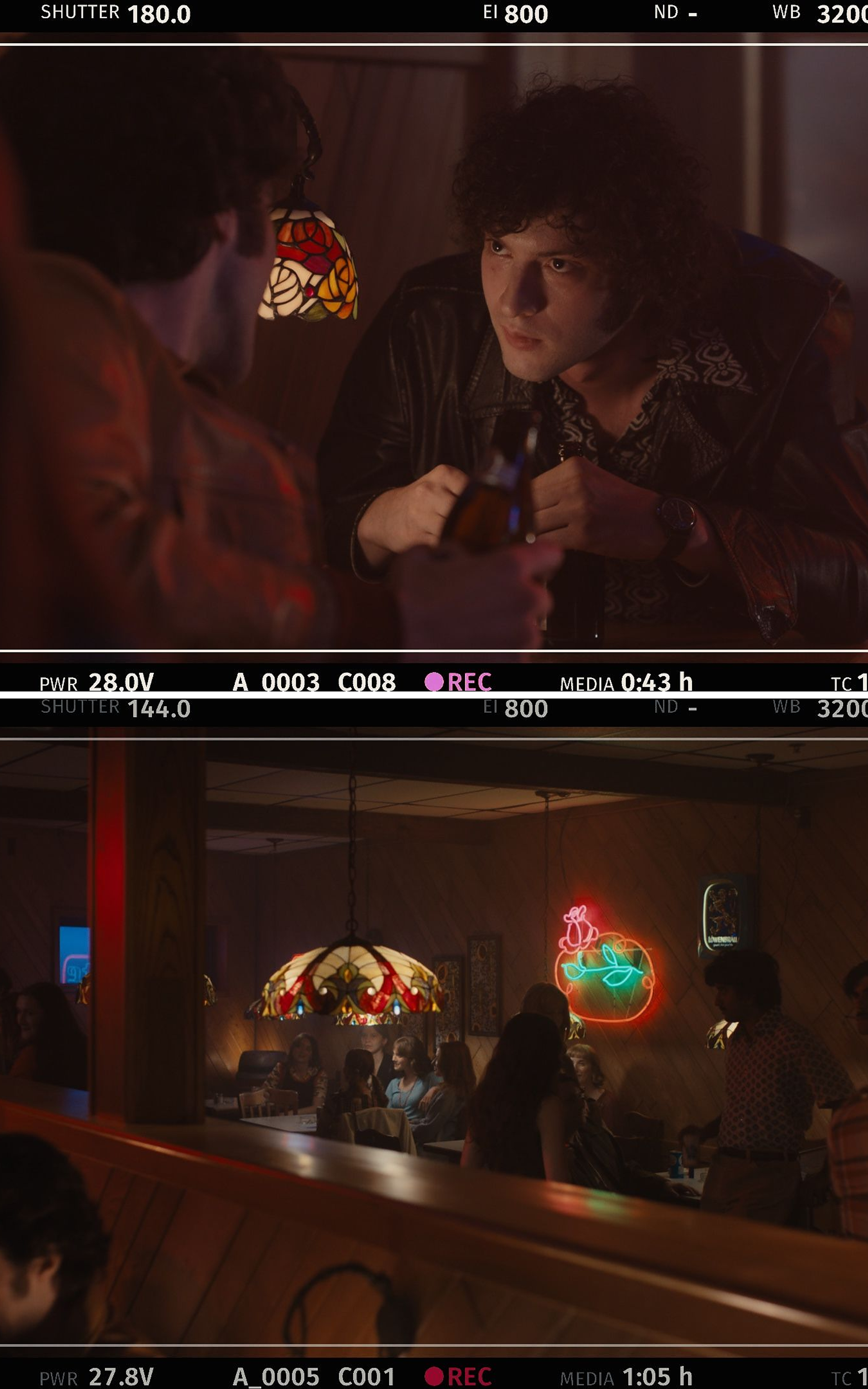 Behind-the-scenes still from a camera monitor showing a curly-haired man in a leather jacket holding a beer bottle across a table from another man seen from behind, lit by a stained-glass lamp; Behind-the-scenes still from a camera monitor showing a dimly lit, retro-style bar scene: patrons seated at wood-paneled booths under a stained-glass pendant lamp, with neon beer and rose signs glowing on the wall