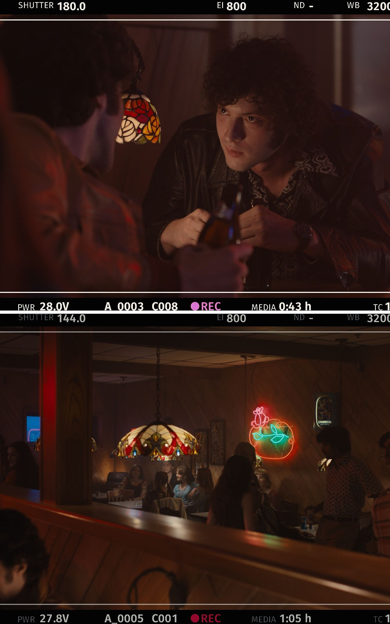 Behind-the-scenes still from a camera monitor showing a curly-haired man in a leather jacket holding a beer bottle across a table from another man seen from behind, lit by a stained-glass lamp; Behind-the-scenes still from a camera monitor showing a dimly lit, retro-style bar scene: patrons seated at wood-paneled booths under a stained-glass pendant lamp, with neon beer and rose signs glowing on the wall