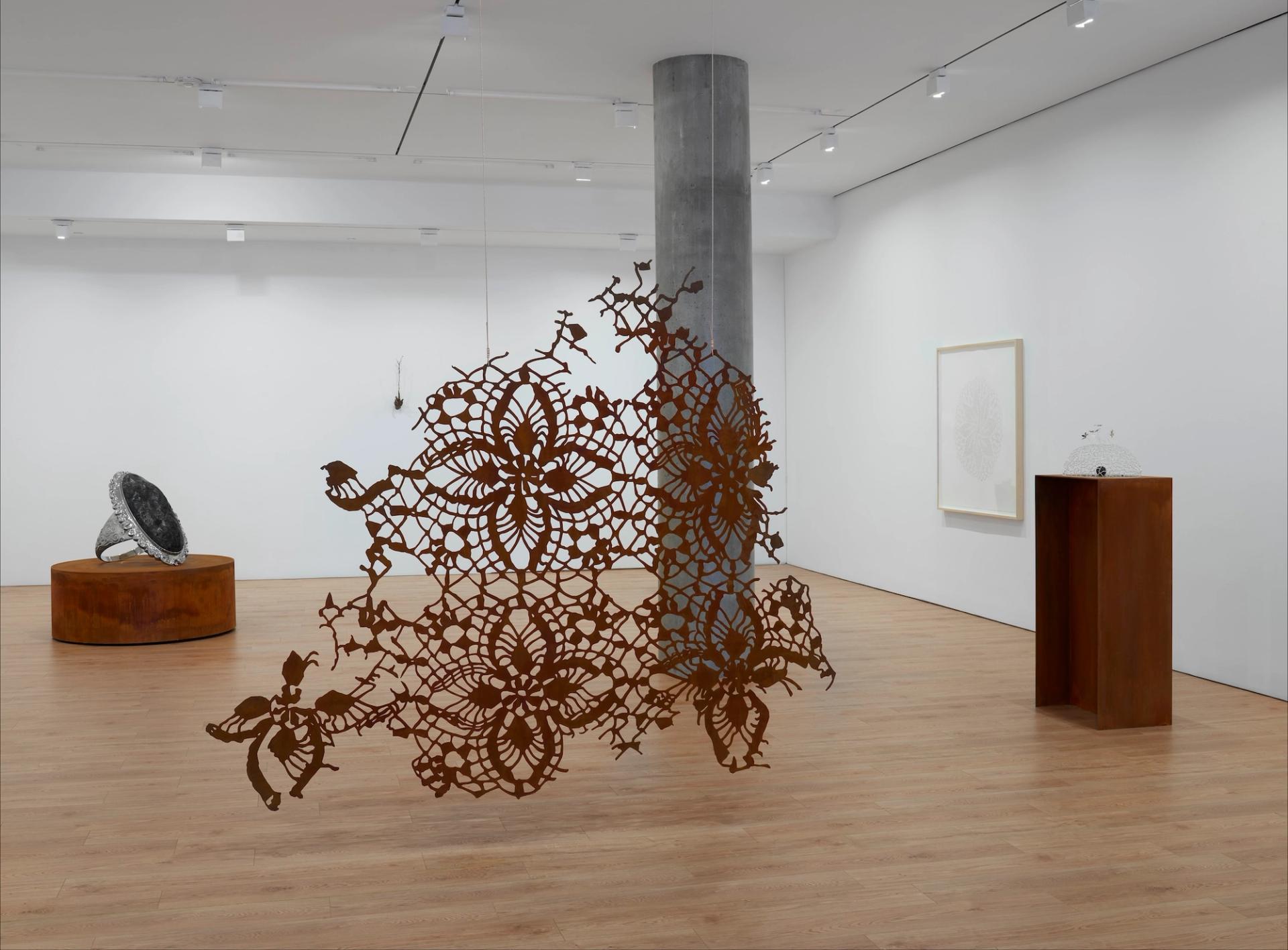 A gallery installation featuring a large rust-colored steel lace sculpture wrapping around a concrete column, with an oversized ring sculpture and a framed work visible in the background