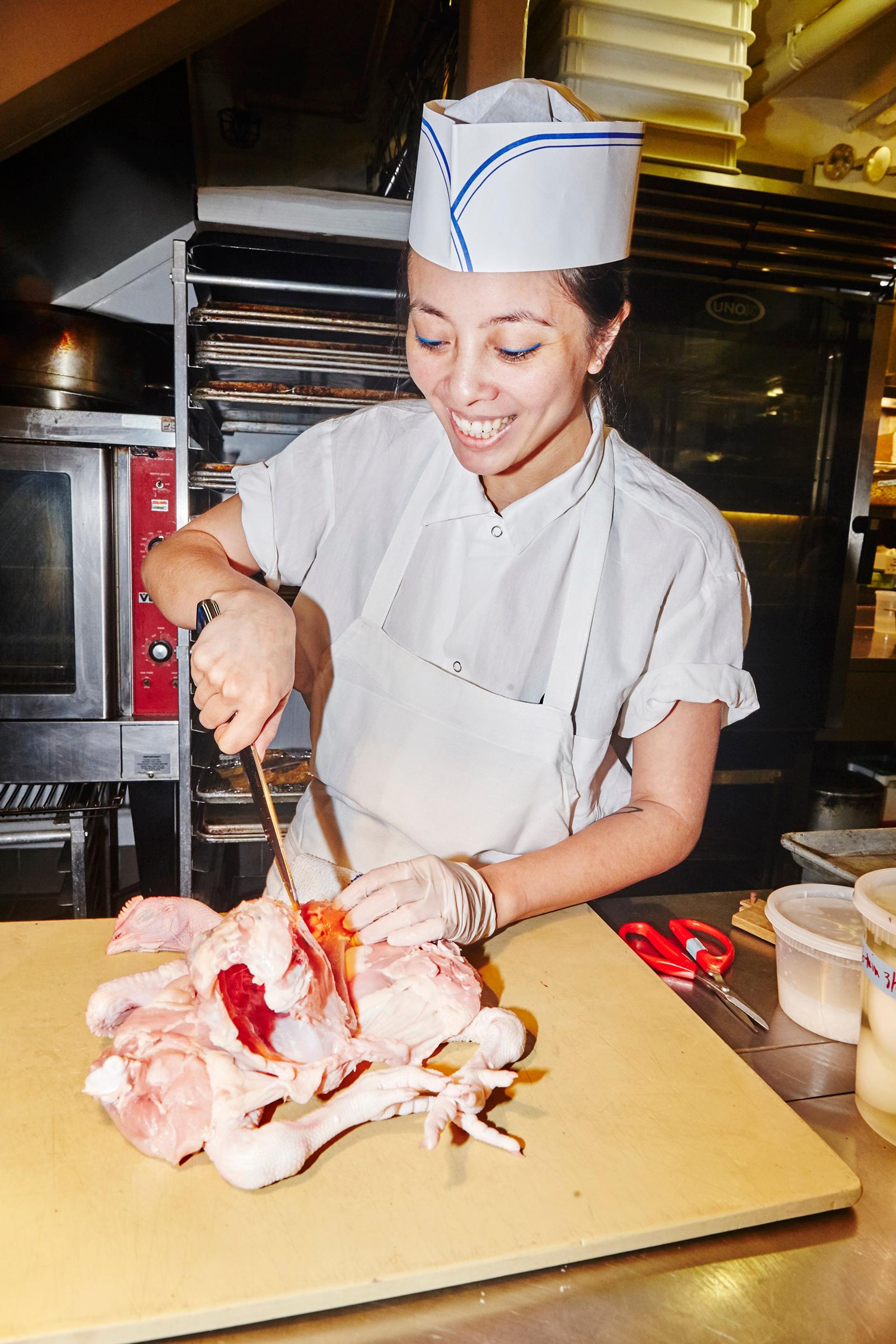 A person in a white apron and chef’s hat butchers a chicken on top of a large cutting board.