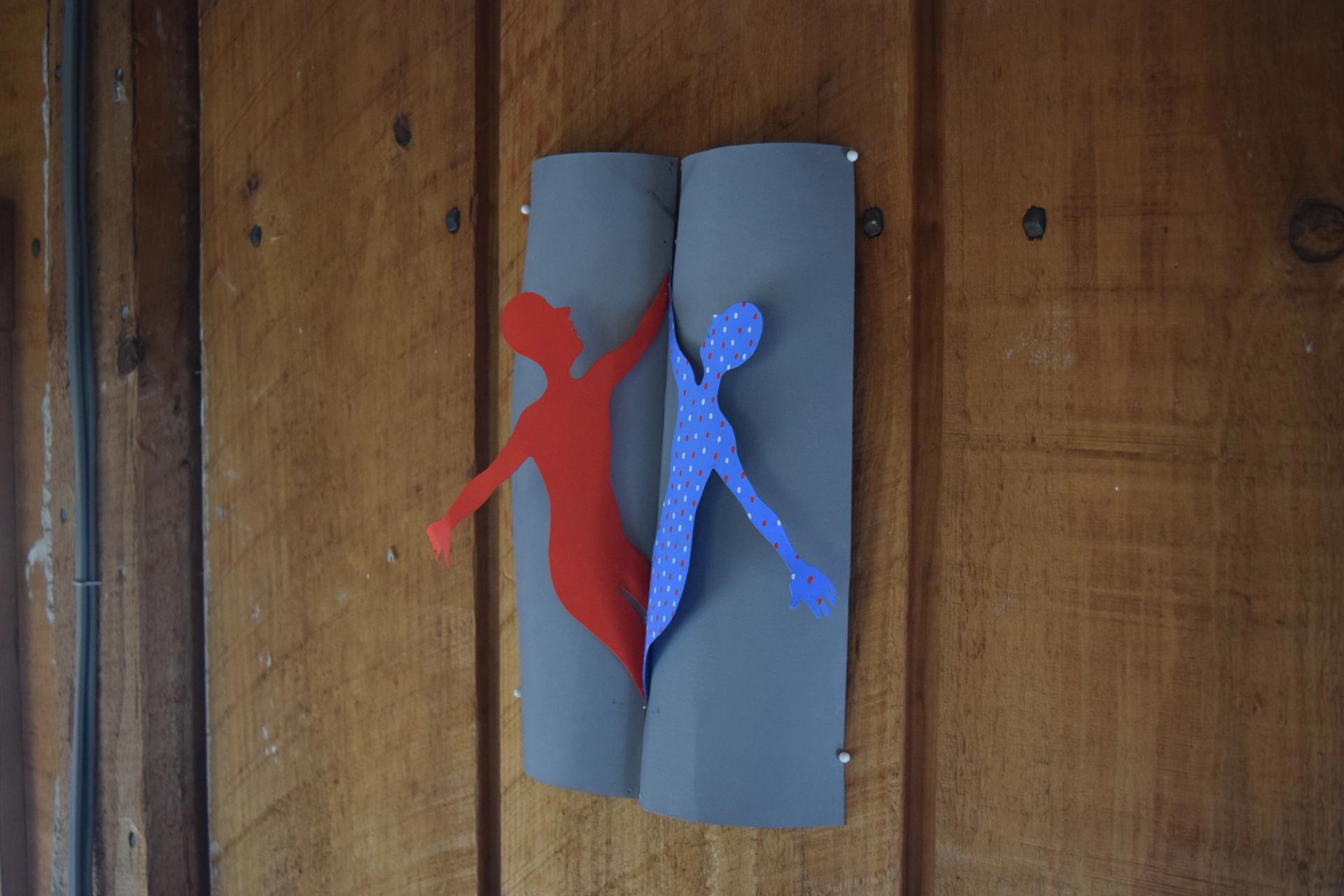 A paper cutout artwork pinned to a wooden wall, depicting two intertwined human silhouettes — one red and one blue with a polka-dot pattern — on a folded gray background.