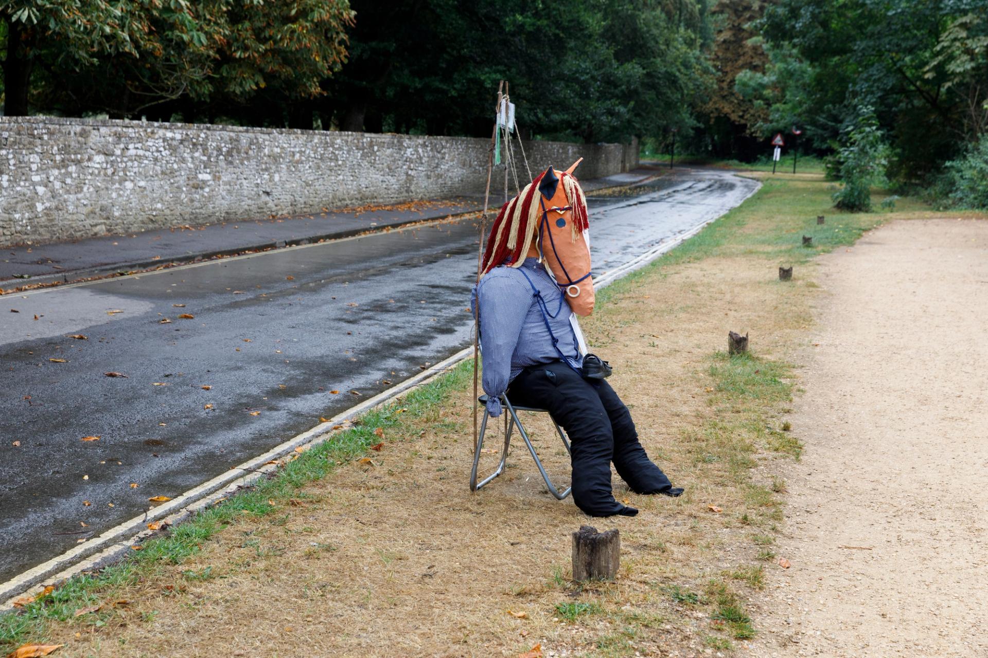 A scarecrow with a horse head sits on a chair beside a road in the grass.