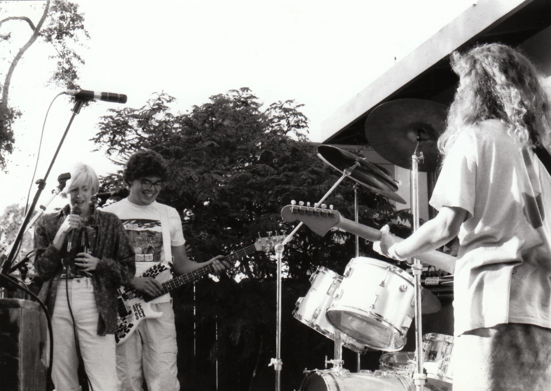 A black and white photograph of three musicians performing outdoors, with a vocalist at a microphone, a guitarist, and a bassist.