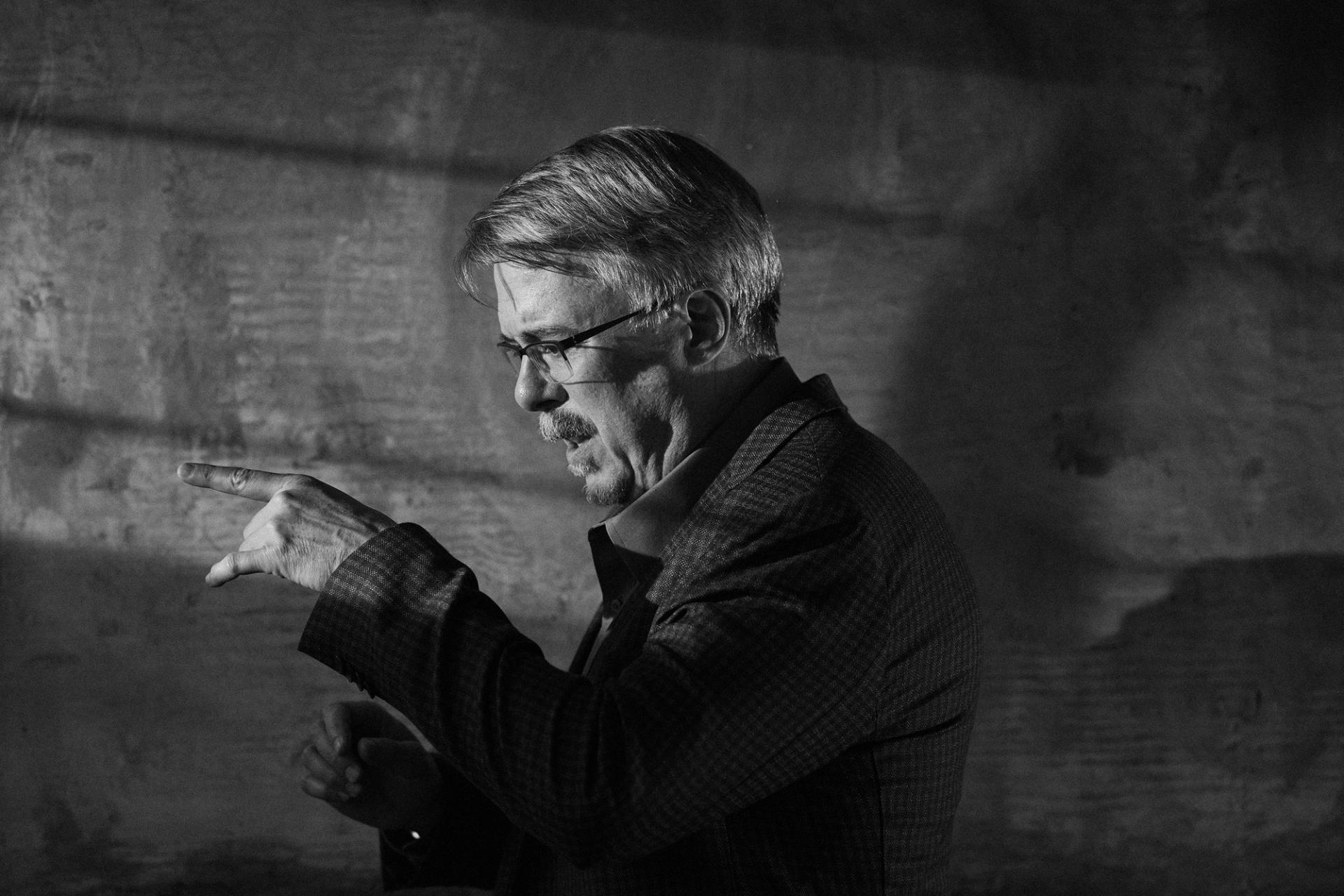 Black-and-white photo of a man in a plaid blazer gesturing with a raised, pointing hand as he speaks, lit dramatically against a textured wall
