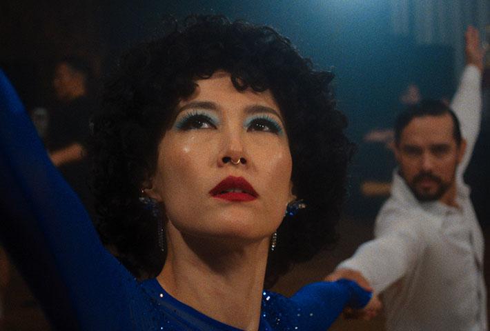 Color film still of a woman with curly dark hair, bold blue eyeshadow, and red lipstick in a sparkling blue dress, her arm raised mid-dance move, with a man in a white shirt striking a matching pose behind her