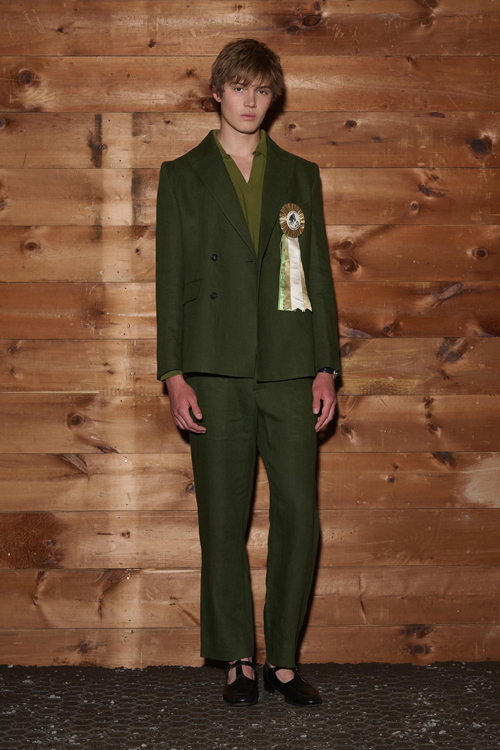 A model stands against a wood-paneled wall wearing a forest green double-breasted suit with a matching shirt, adorned with a decorative rosette ribbon on the lapel