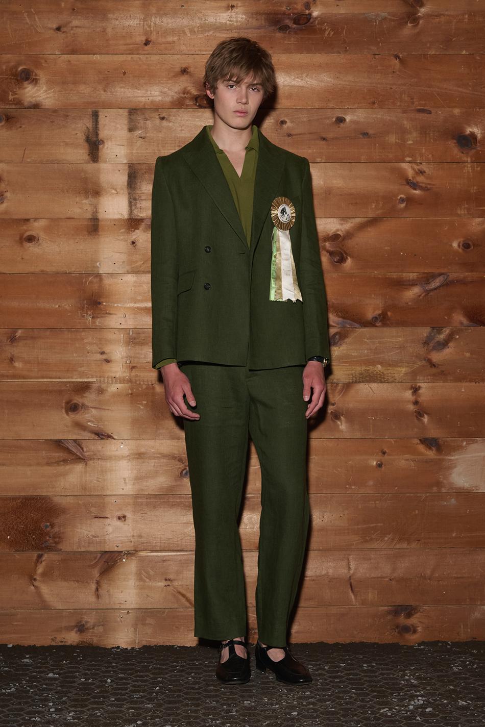 A model stands against a wood-paneled wall wearing a forest green double-breasted suit with a matching shirt, adorned with a decorative rosette ribbon on the lapel