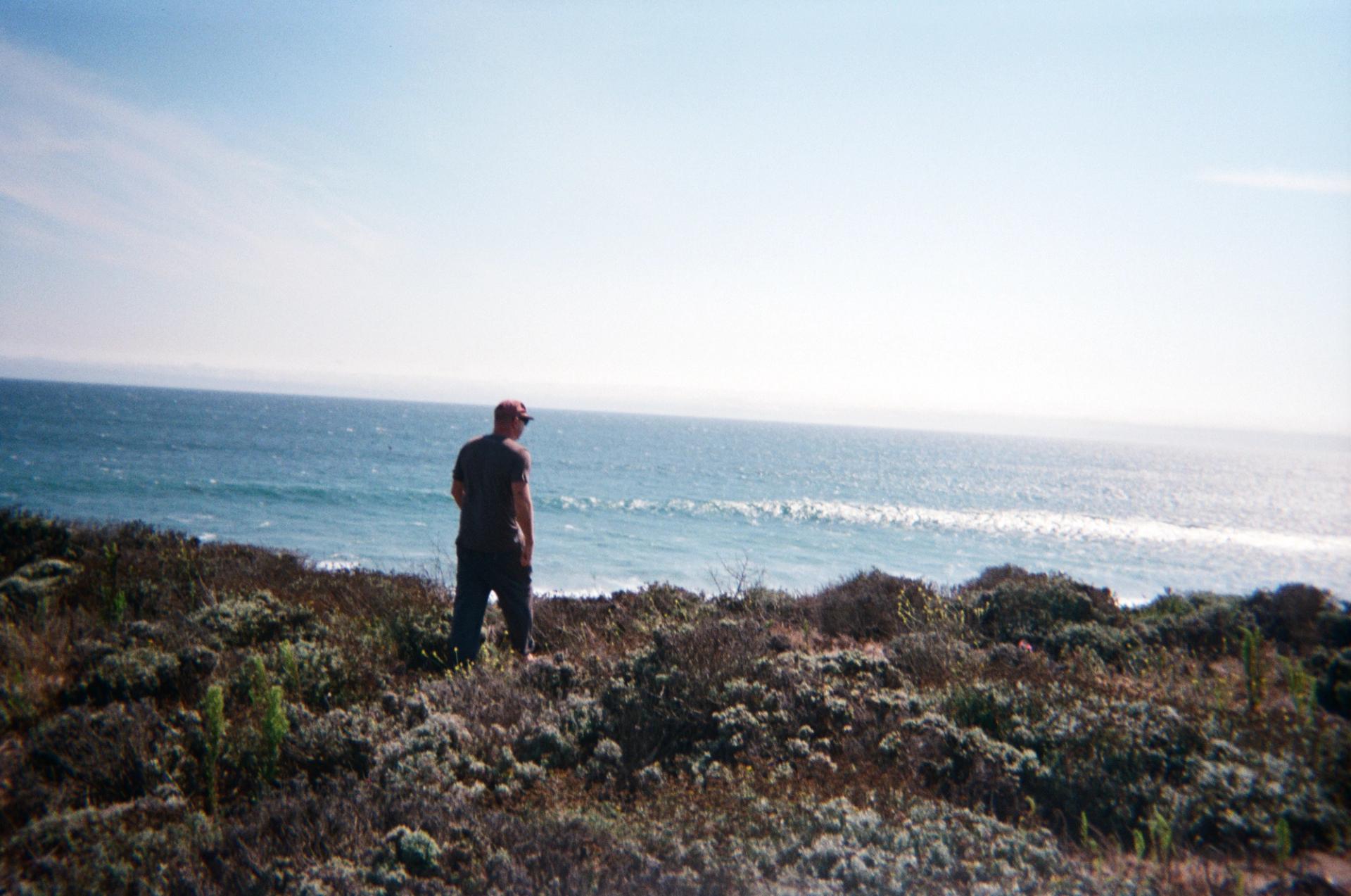 A man in a cap standing among coastal brush atop a bluff, looking out at the ocean with waves breaking below under a hazy sky.