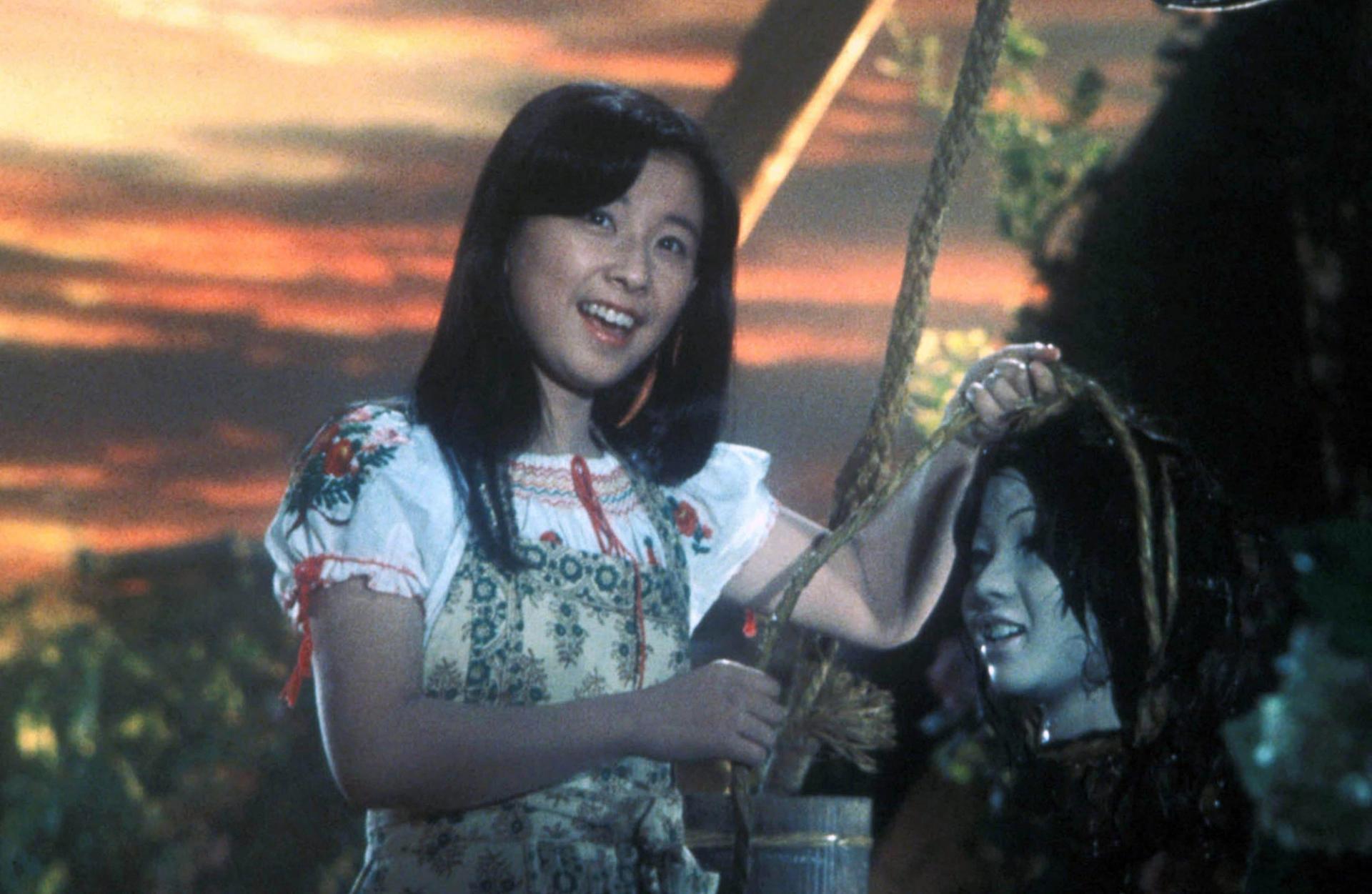 A young girl smiles while holding a smiling severed head outdoors.