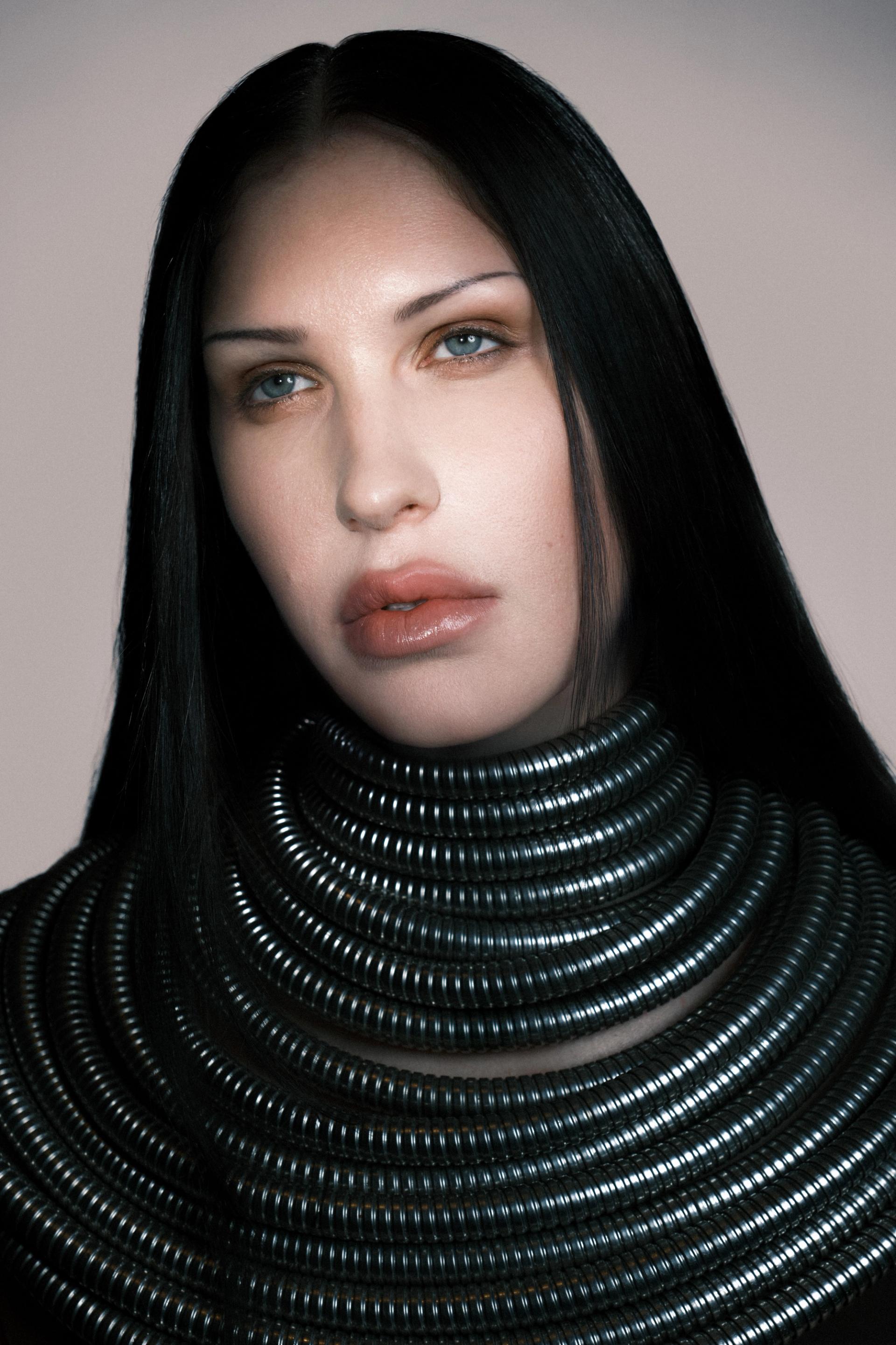 Studio portrait of a person with long black hair wearing layered black coiled tubing around the neck and shoulders, gazing directly at the camera against a plain background.