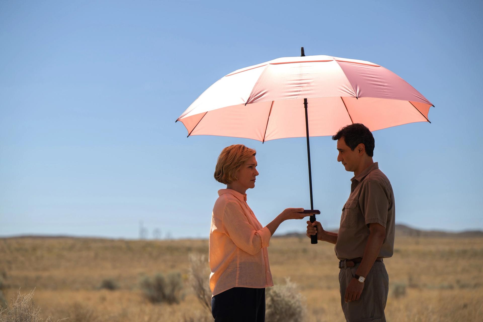 A woman and man standing face to face under a large pink-and-white umbrella in a dry, open landscape, both gripping its handle between them