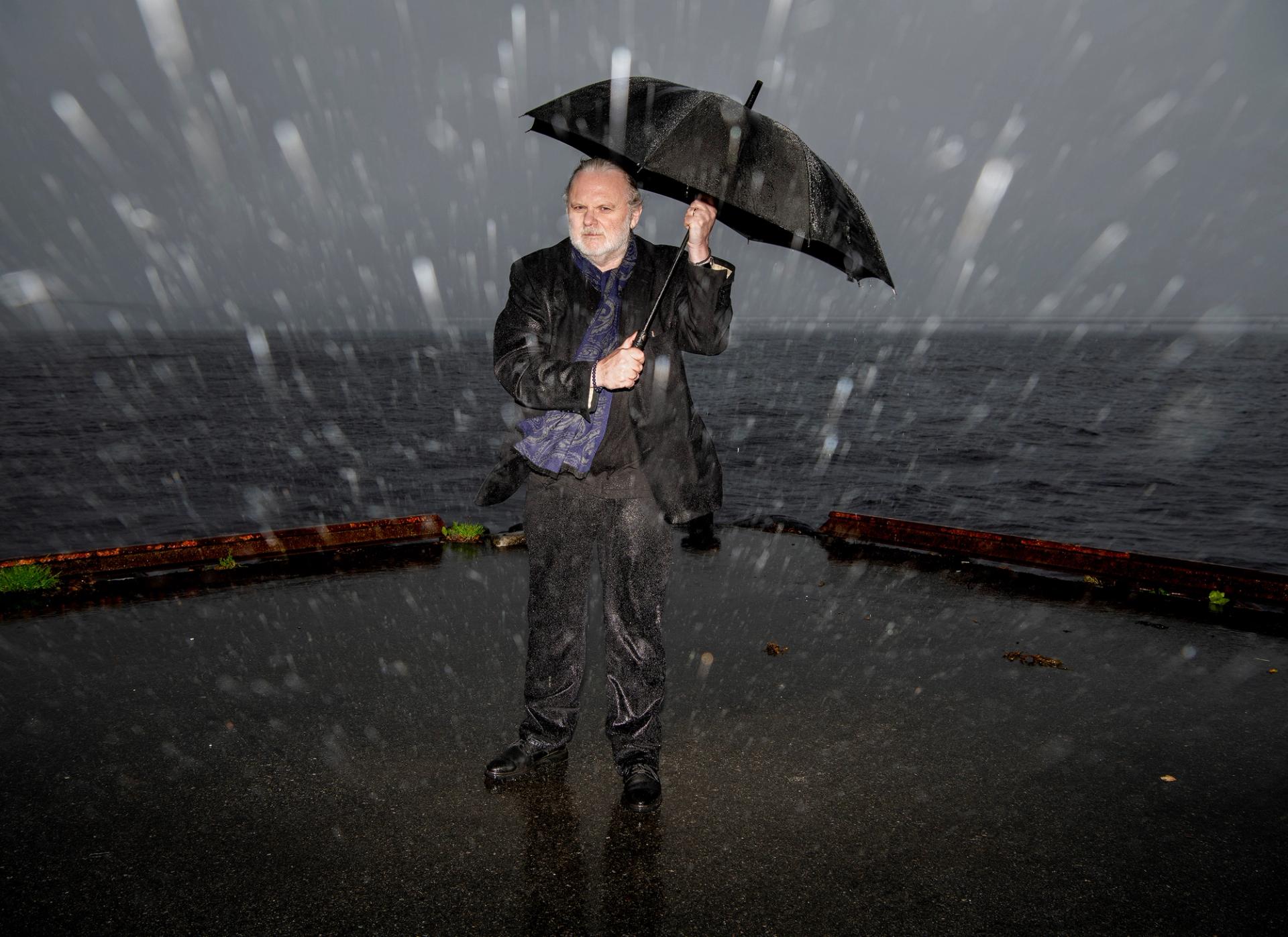 Man with a white beard stands outside holding a black umbrella in heavy rain, with the grey ocean behind him.