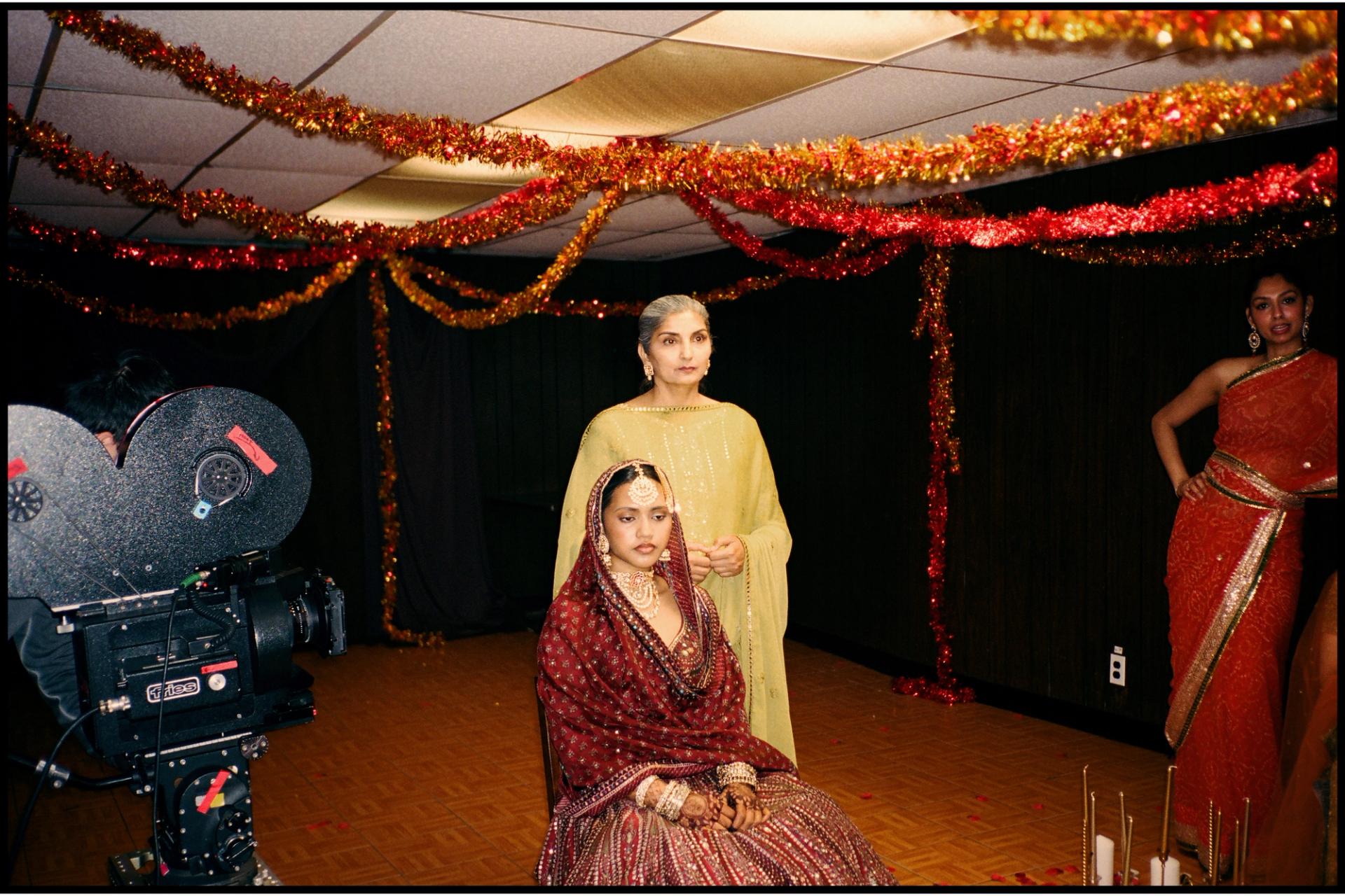 A film set decorated with red and gold tinsel garlands, with a bridal figure in burgundy seated before a film camera while two other performers in South Asian attire stand nearby