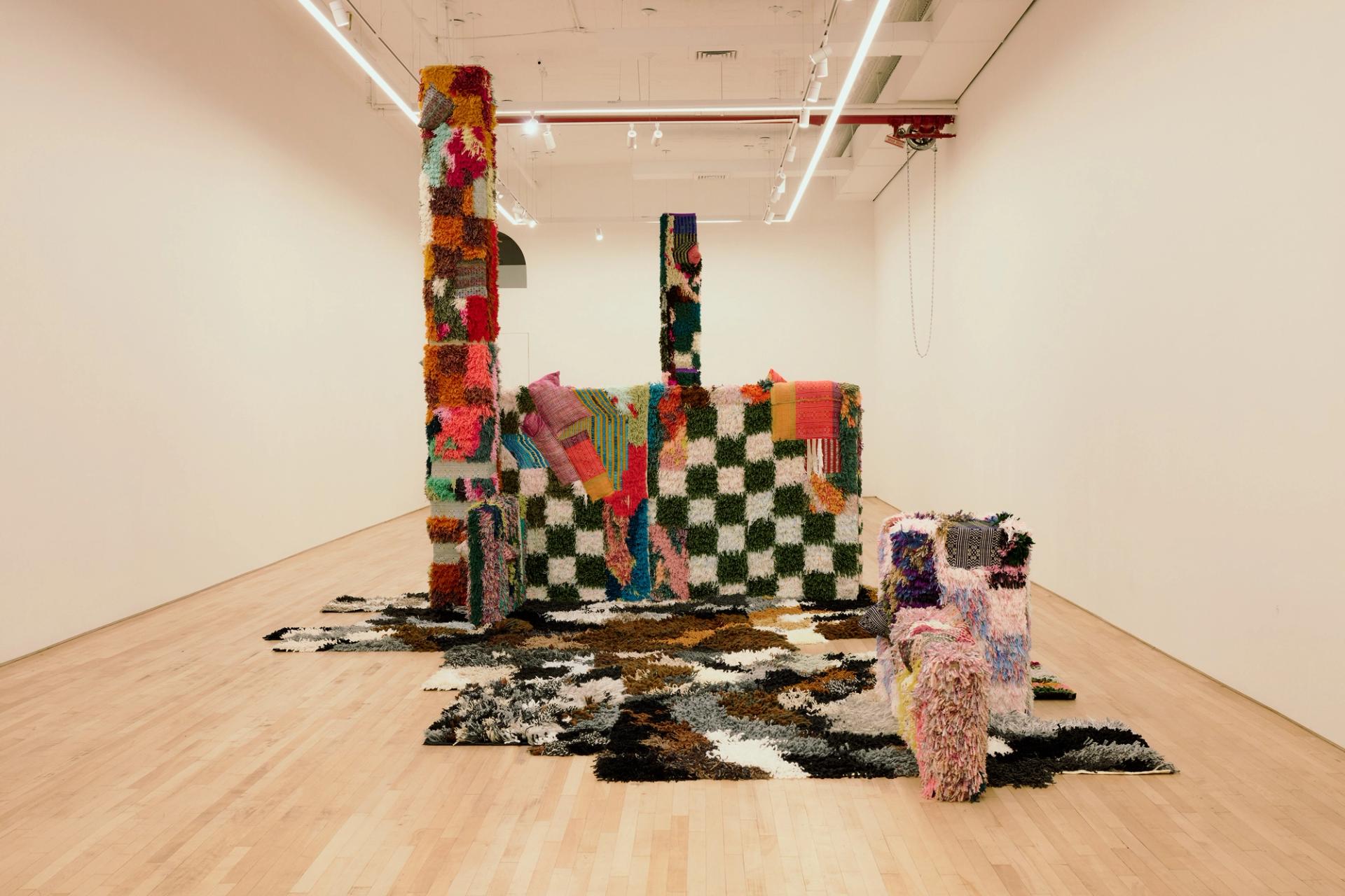 Textile art installation in a white gallery: tall shaggy columns and a boxy, checkerboard-patterned structure covered in colorful woven and tufted fabric, with matching patchwork rugs spilling across the wooden floor.