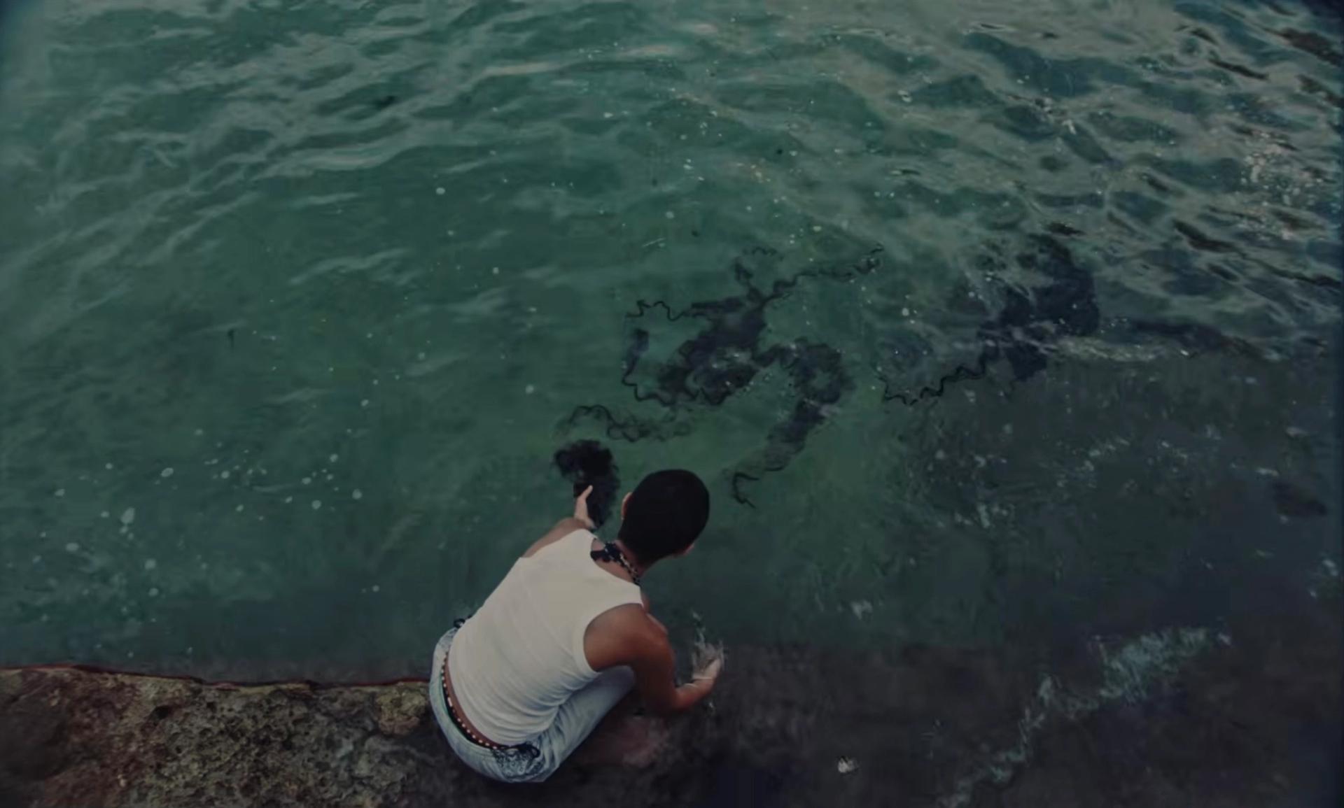 Overhead view of a person crouching on rocks, touching the water beside drifting seaweed.