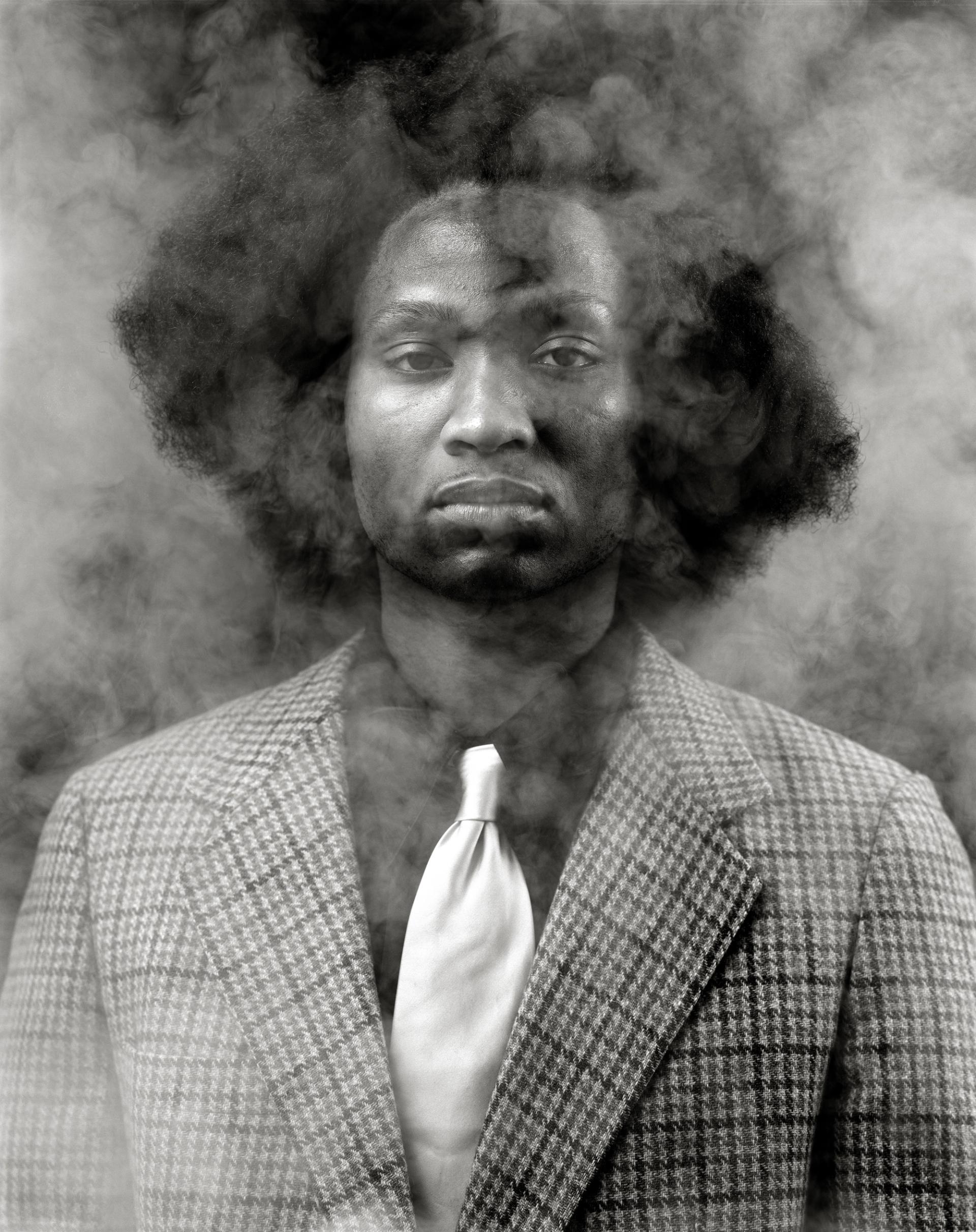 Black-and-white portrait of man in checked suit and tie, surrounded by swirling smoke.