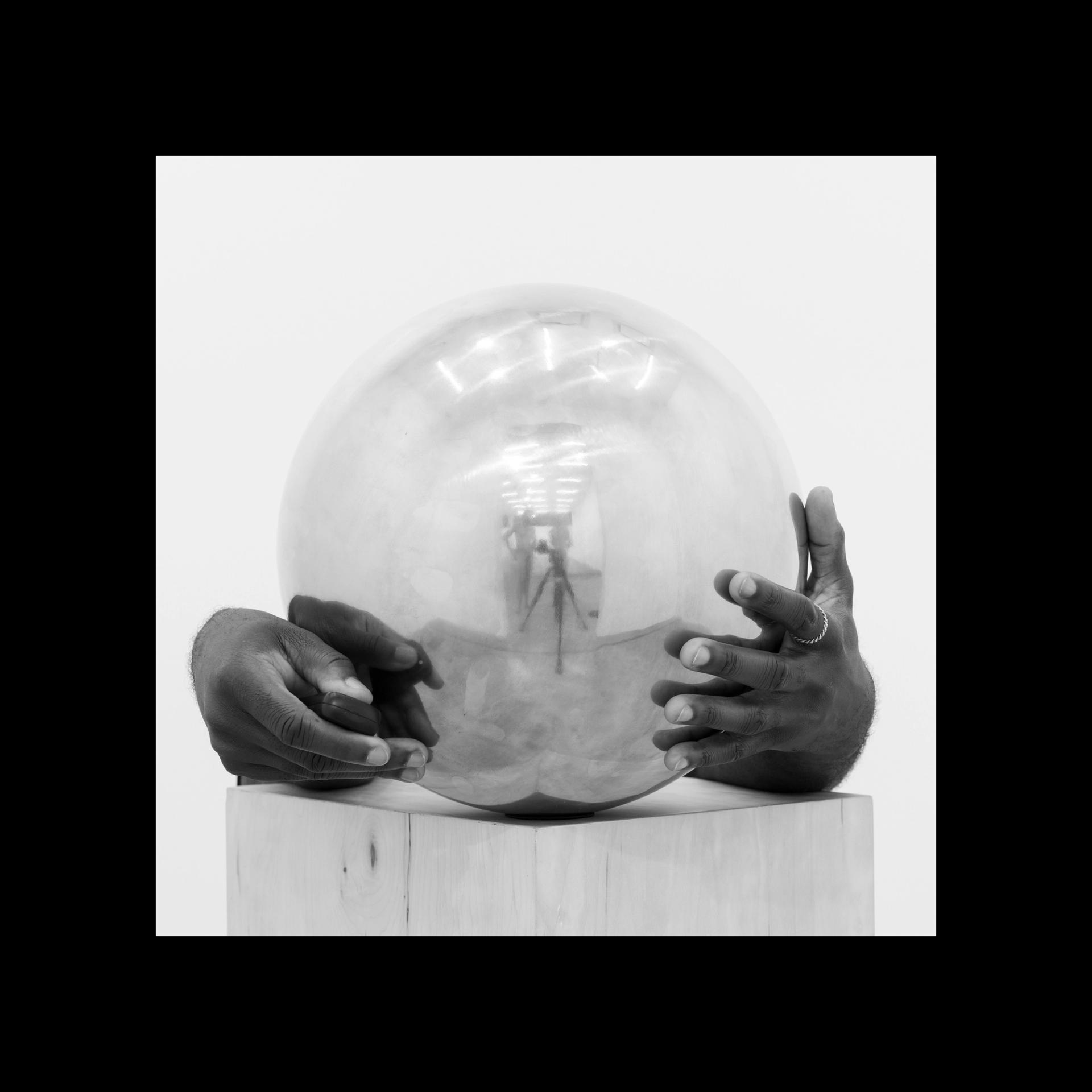 Hands cradle a reflective silver sphere that mirrors a gallery space and camera tripod.