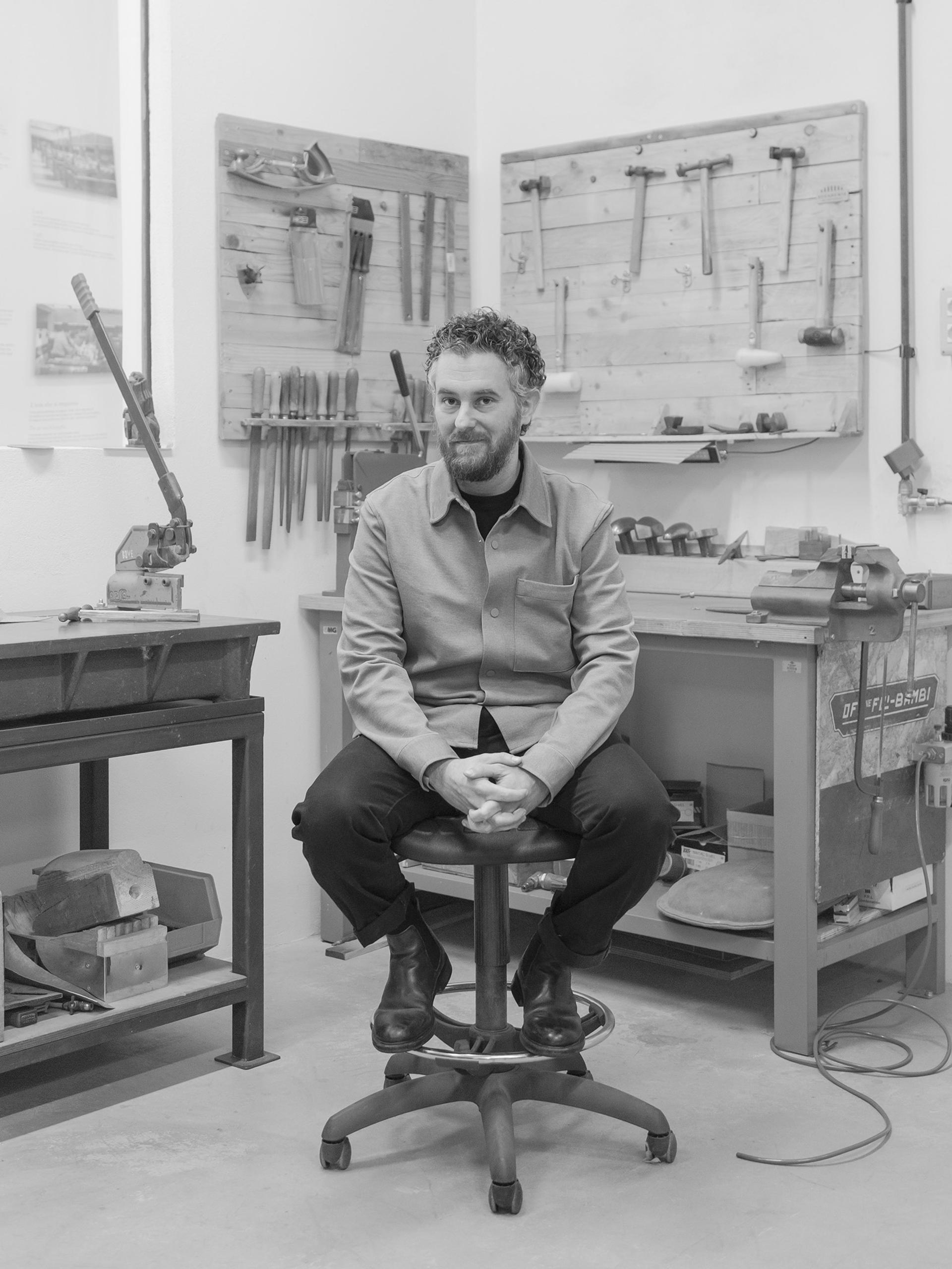 Black-and-white photograph of a man sitting on a stool in a workshop.